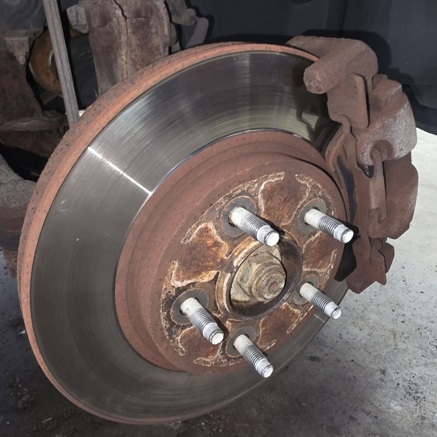 Close-up of a car brake rotor and caliper with rusted lug studs on a wheel hub.