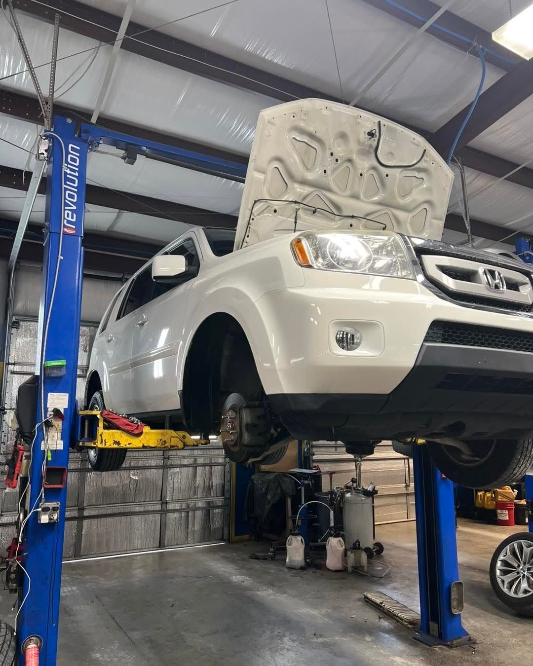 White SUV lifted in an auto repair shop with its hood open on a blue car lift.