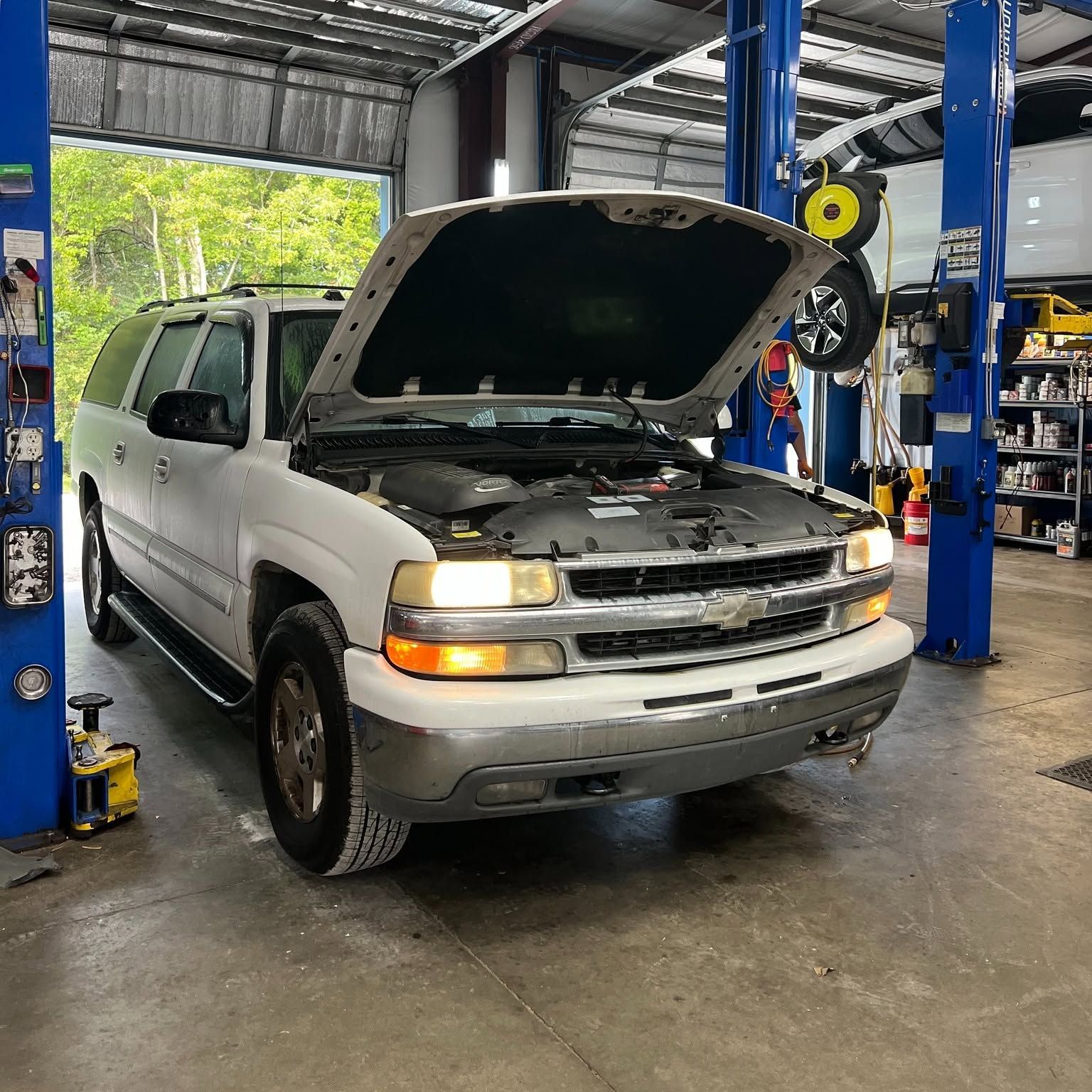White SUV with hood open in an auto repair shop, under a lift with tools and tires nearby