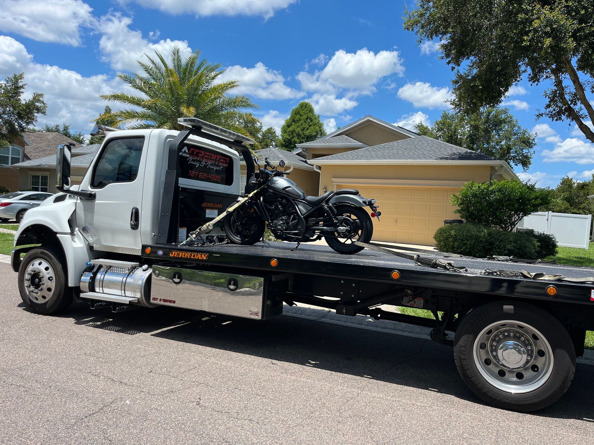 White tow truck carrying a black motorcycle on a sunny street in front of a house.