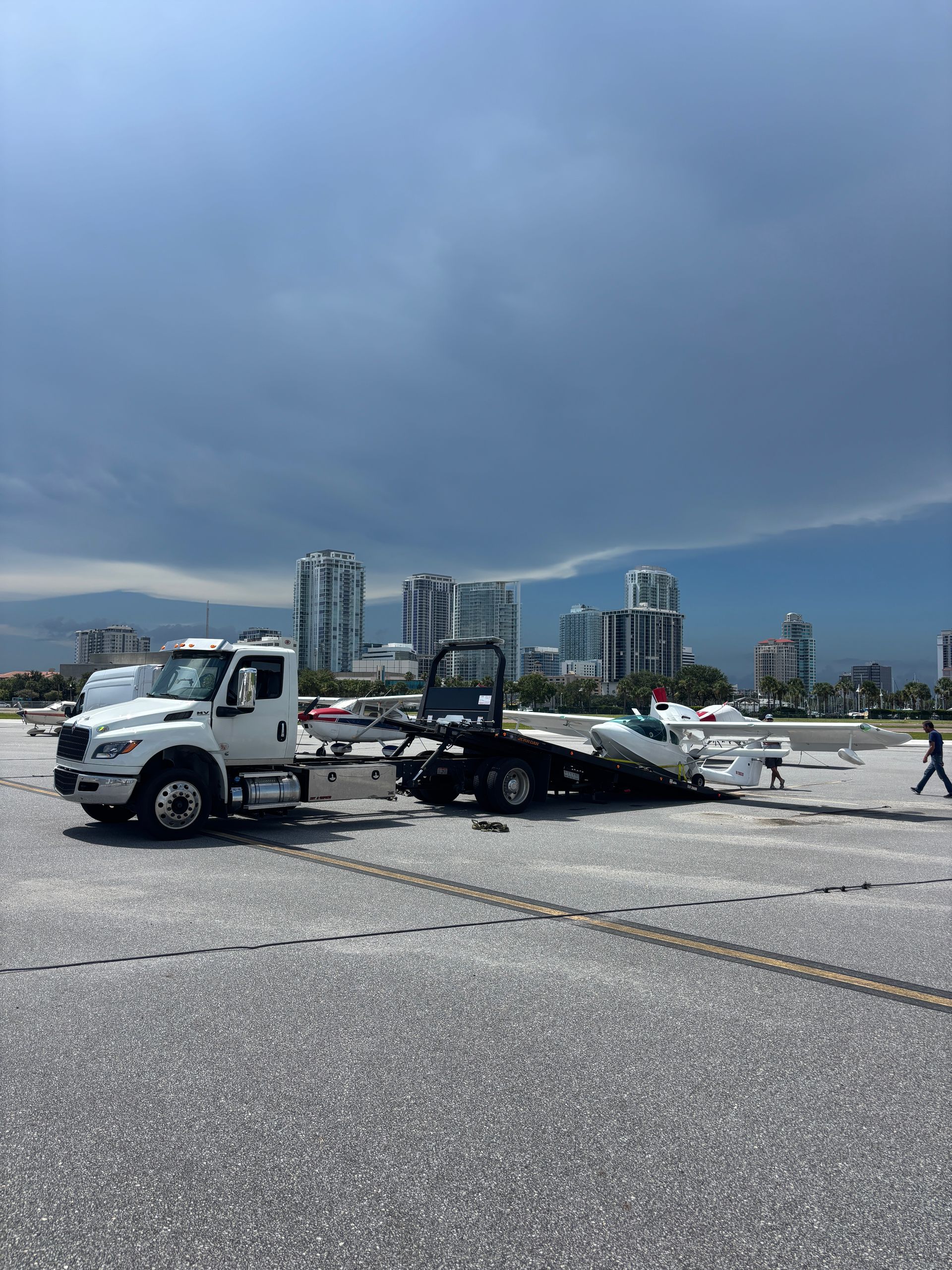A white tow truck pulling a boat on a trailer across an asphalt surface, city buildings in the background.