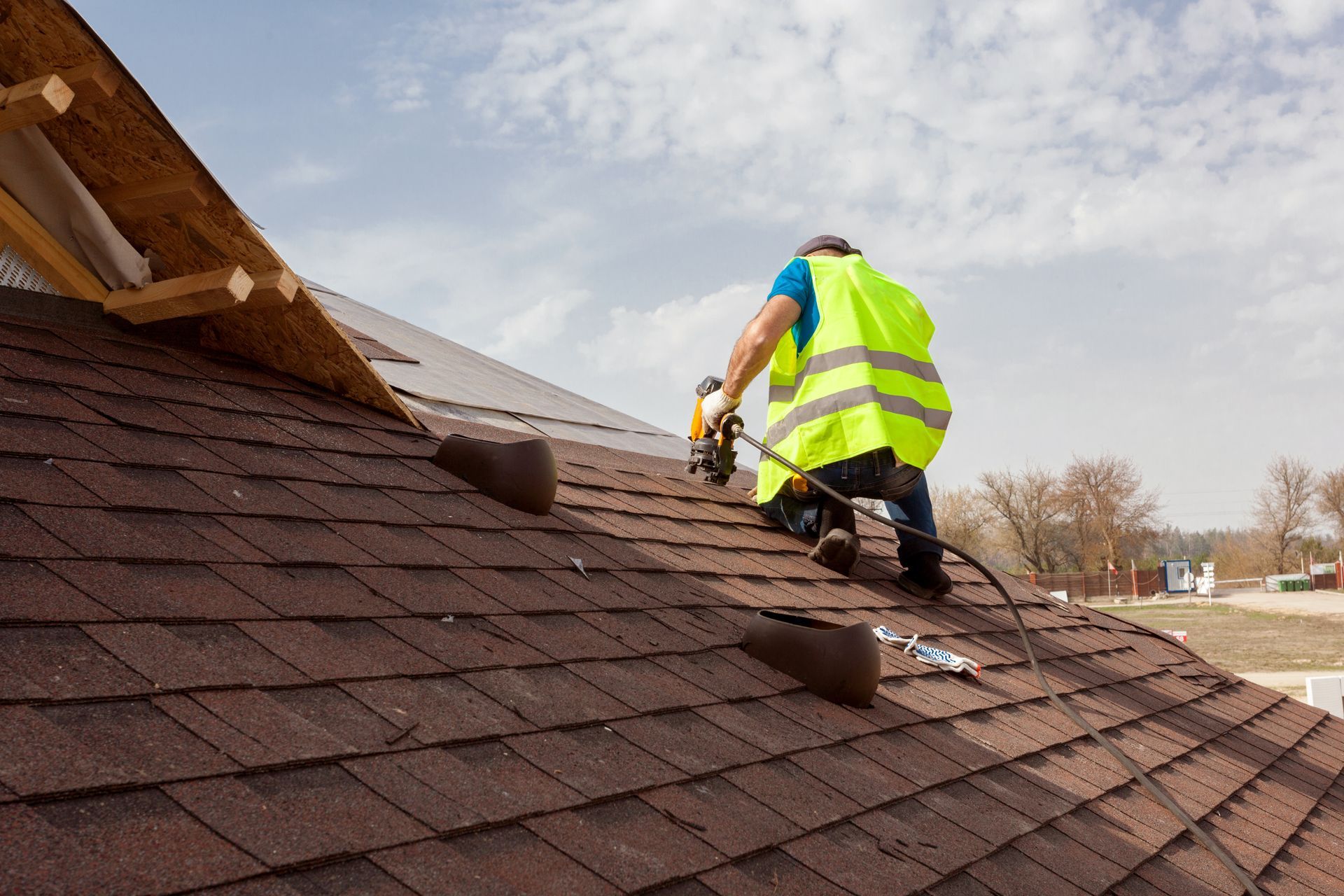 Roofer wearing a safety vest using tools to install asphalt shingles on a sloped house roof. Roofer wearing a safety vest using tools to install asphalt shingles on a sloped house roof.