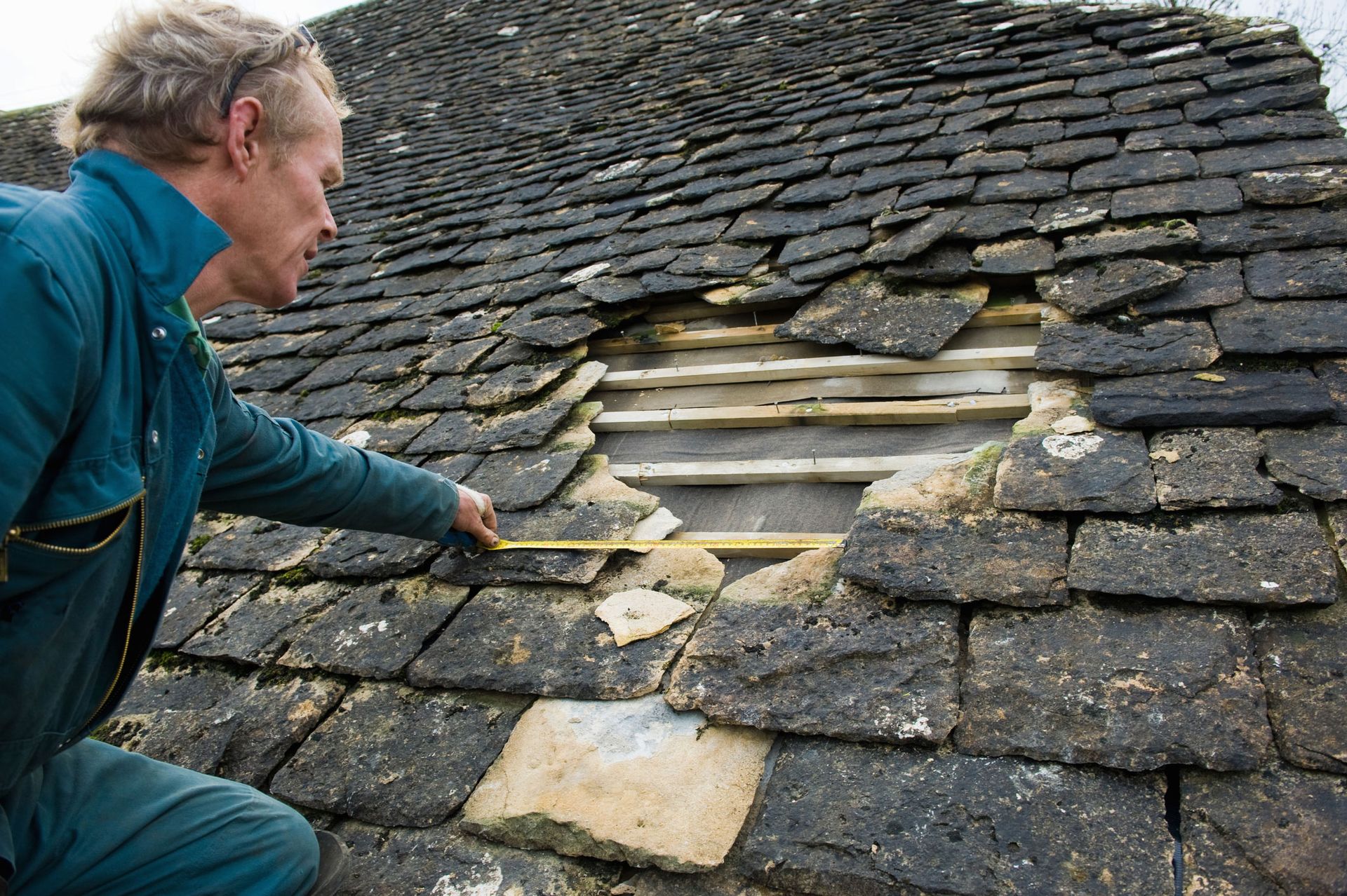 A worker in a blue jumpsuit examines a large hole in a slate roof, holding a measuring tape to the damaged section. A worker in a blue jumpsuit examines a large hole in a slate roof, holding a measuring tape to the damaged section.