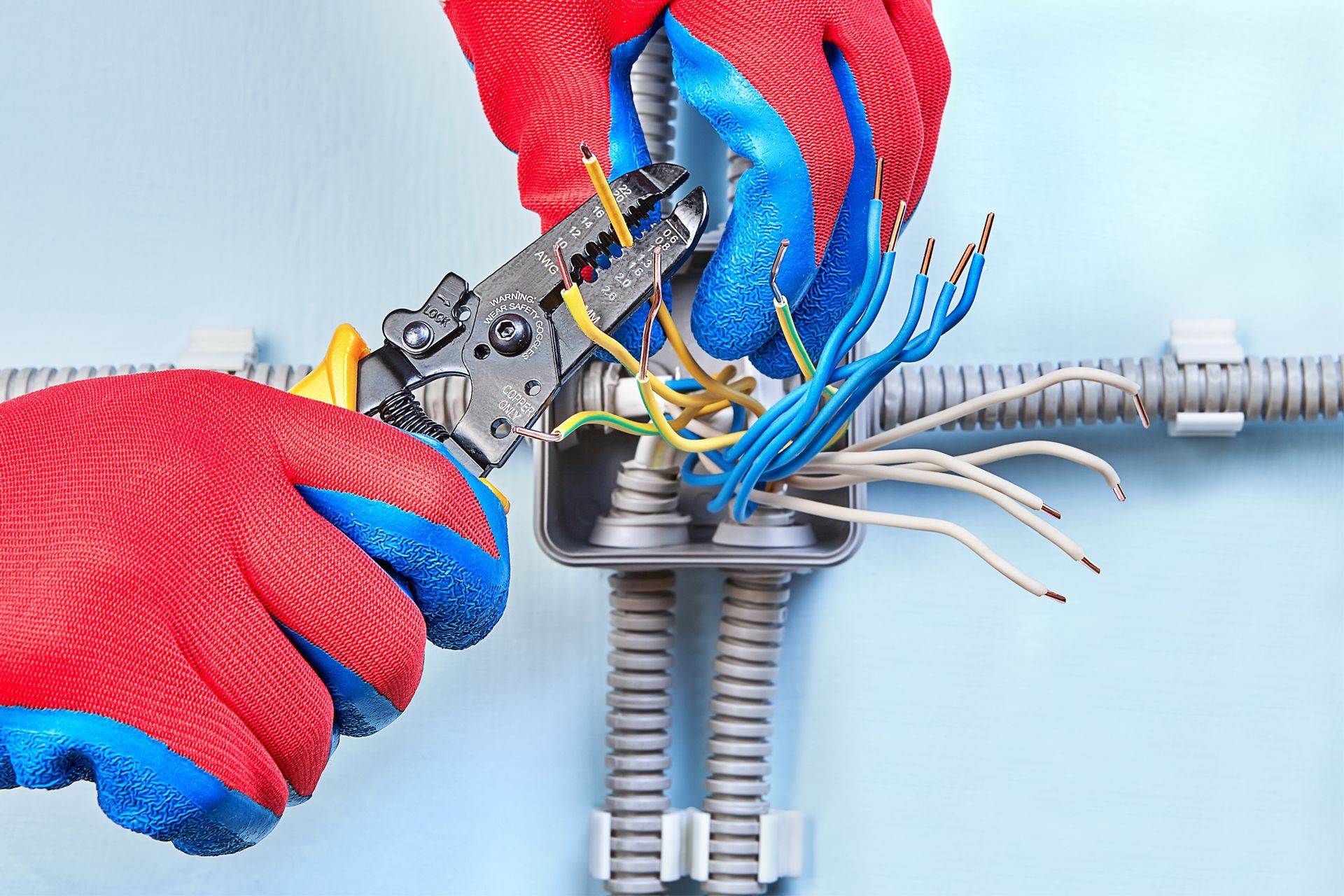 Hands in red gloves strip wires in a junction box, blue and white wires visible on a light blue wall.