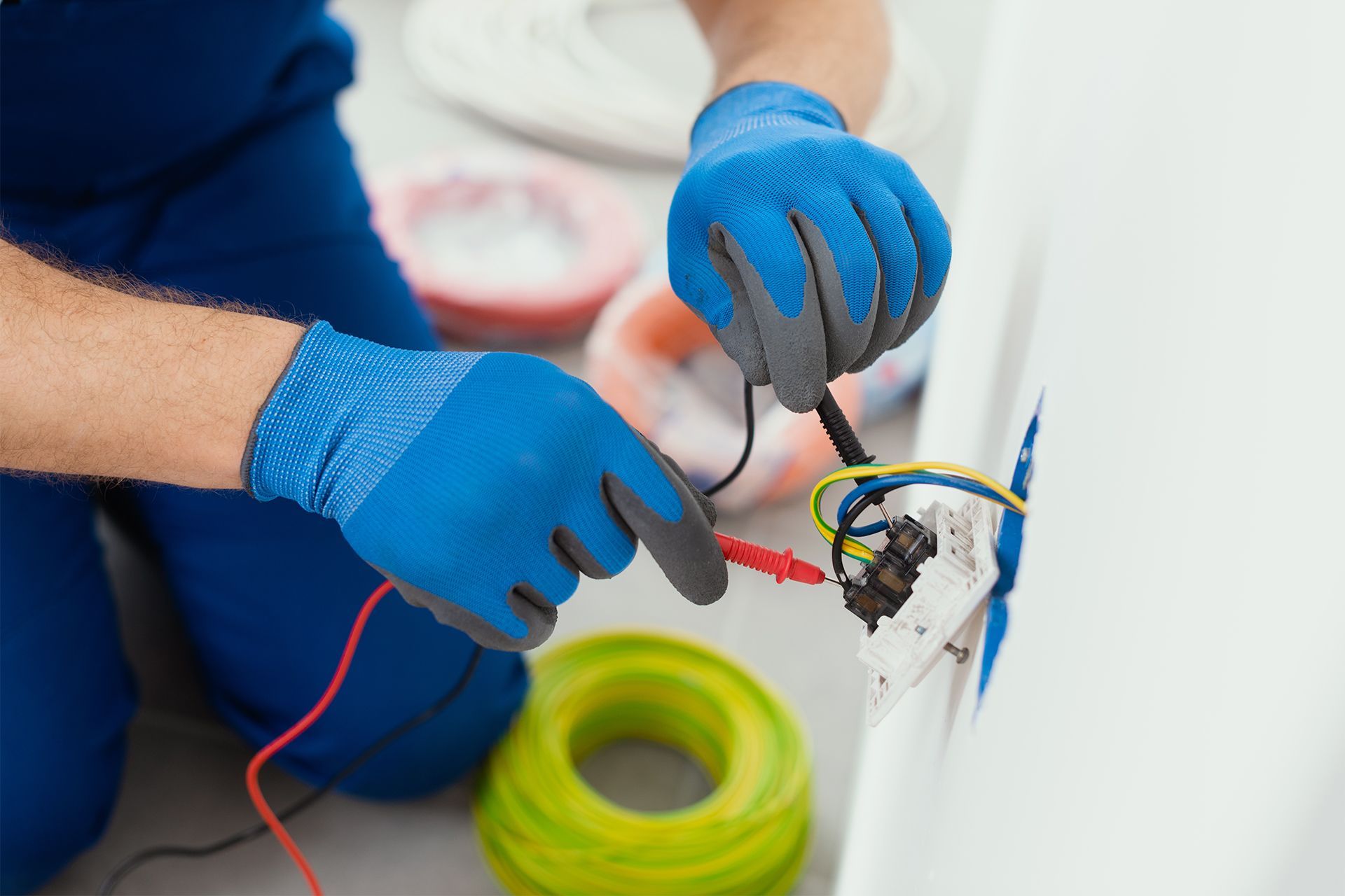 Electrician in blue gloves testing wires in a wall outlet, with spools of colorful wire in the background.