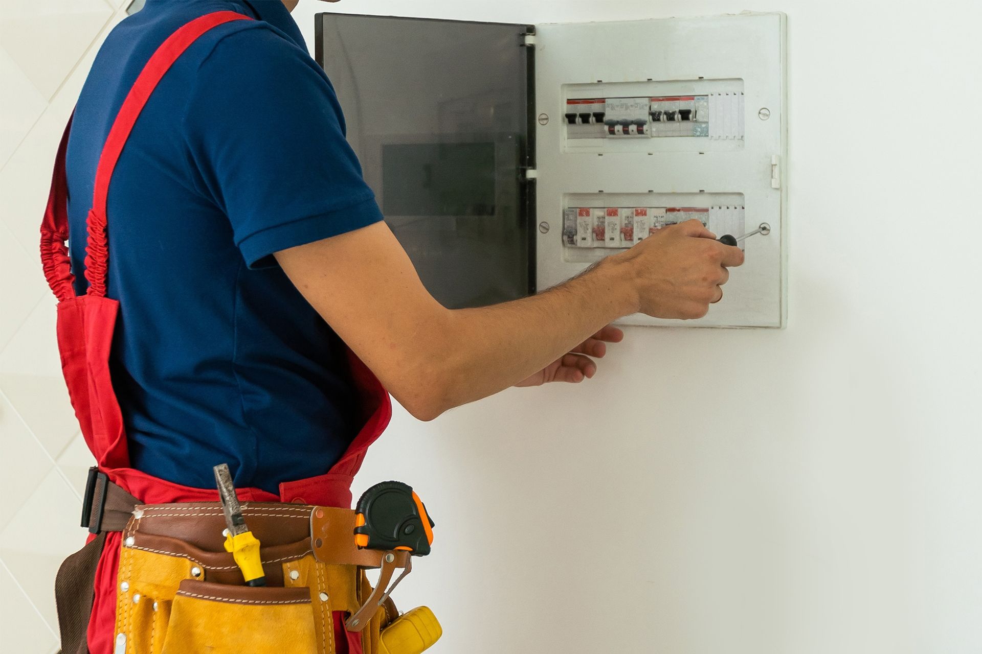 Electrician in blue shirt and red overalls working on a circuit breaker panel.