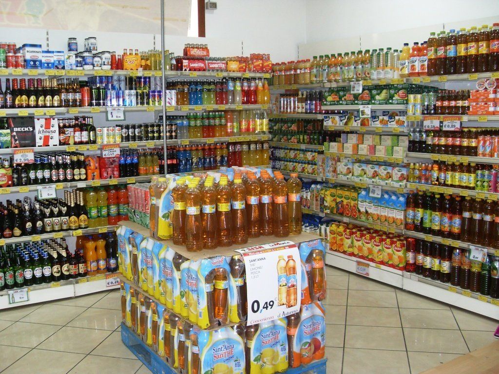Corridoio di un supermercato con scaffali pieni di bevande, bottiglie e prodotti confezionati di vario genere, disposti attorno a un espositore centrale.