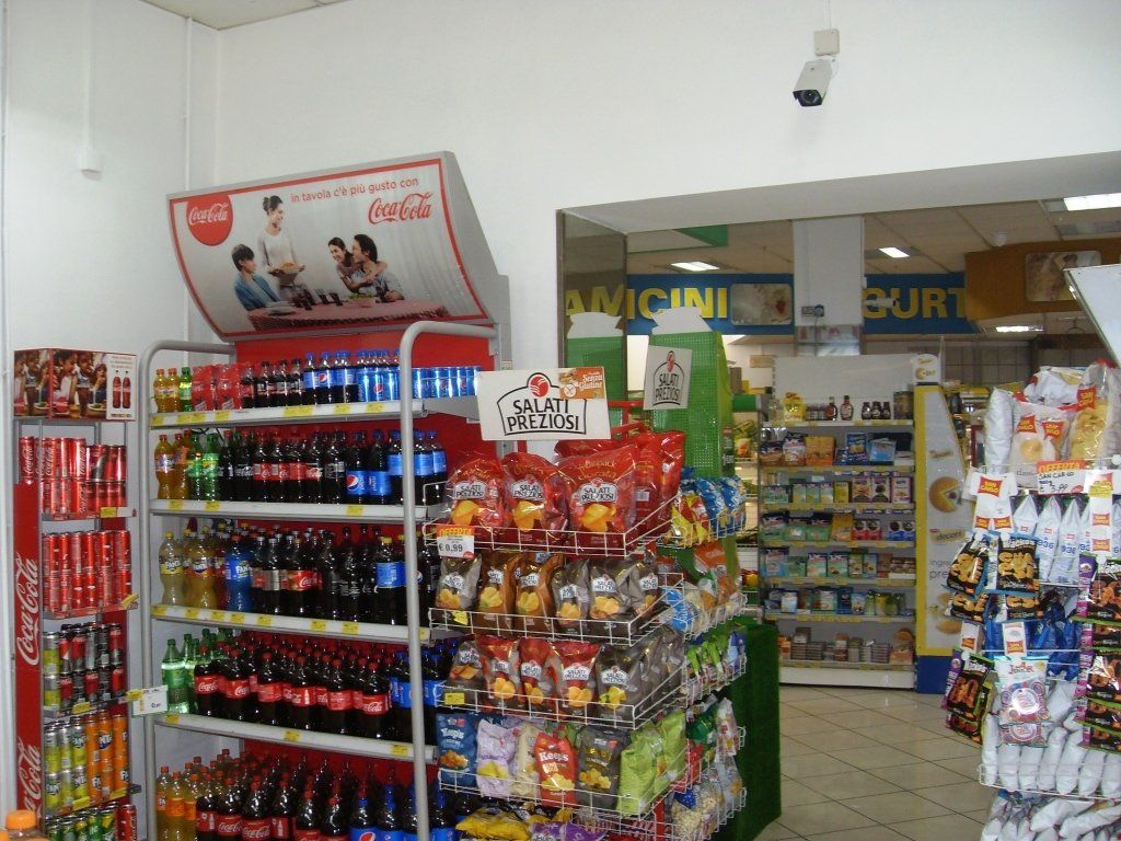 Corridoio del minimarket con snack colorati, scaffali di bibite e un piccolo bancone sul retro.