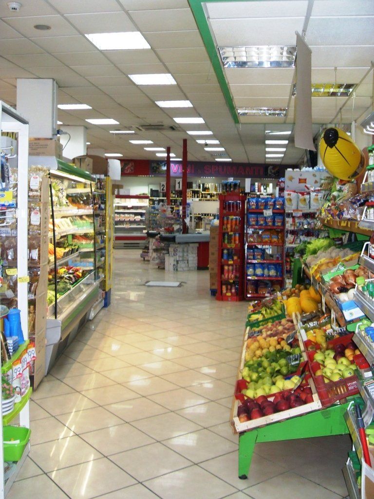 Corridoio di un supermercato con espositori di frutta e verdura, scaffali e illuminazione fluorescente brillante.
