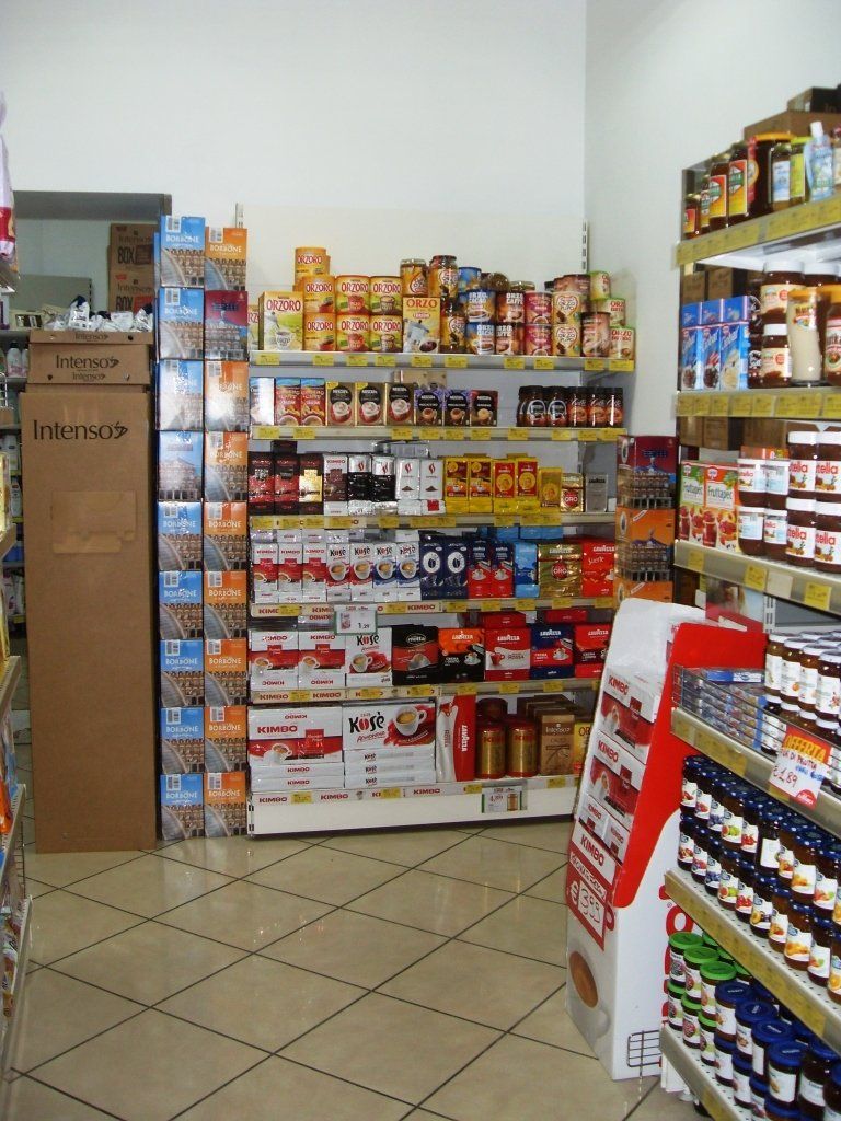 Un piccolo corridoio di un supermercato con scaffali pieni di alimenti confezionati e barattoli, oltre a un espositore di cartone sulla sinistra.