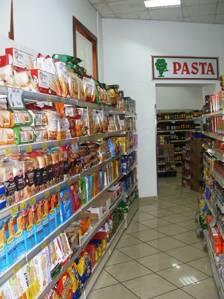 Un corridoio di un supermercato con scaffali pieni di pasta e prodotti confezionati, e un cartello con la scritta 