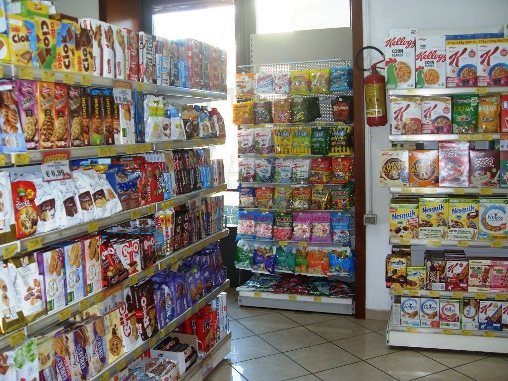 Un piccolo corridoio di un supermercato con snack e cereali confezionati in modo colorato sugli scaffali, illuminato dalla luce naturale.