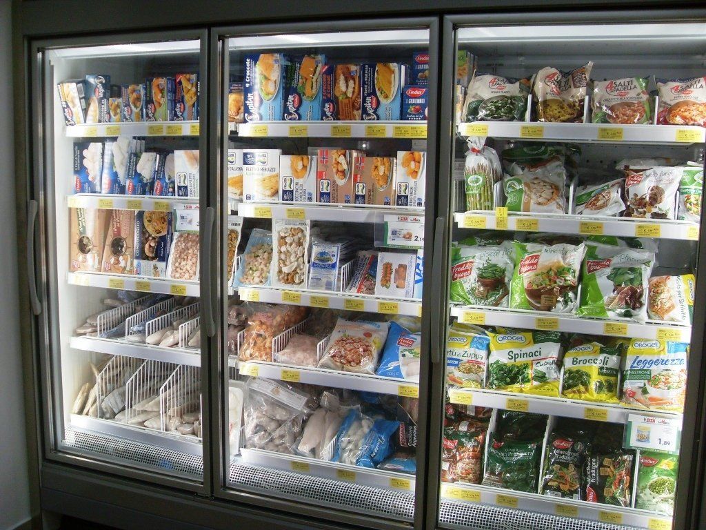 Congelatore aperto per generi alimentari con vari alimenti e bevande confezionati dietro ante di vetro.