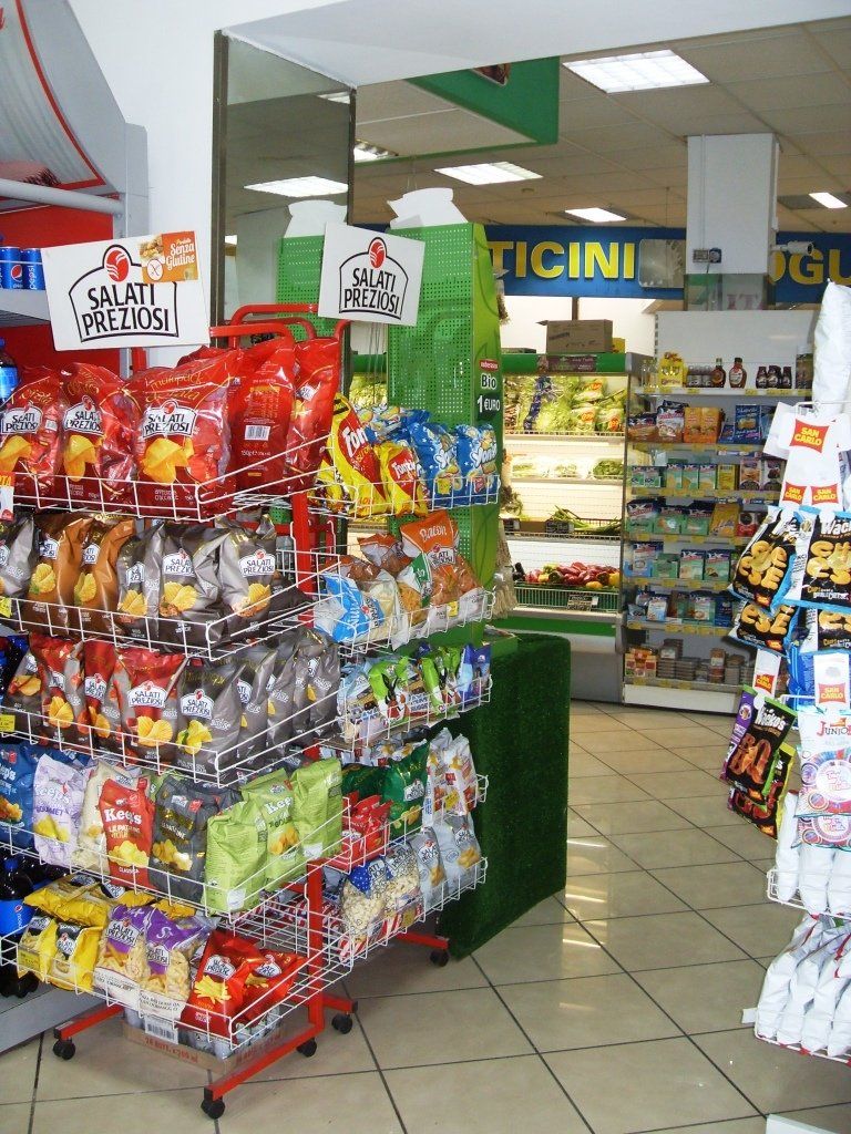 Esposizione di caramelle e snack in un piccolo corridoio di un minimarket, con dolcetti confezionati in modo colorato e pavimento piastrellato.