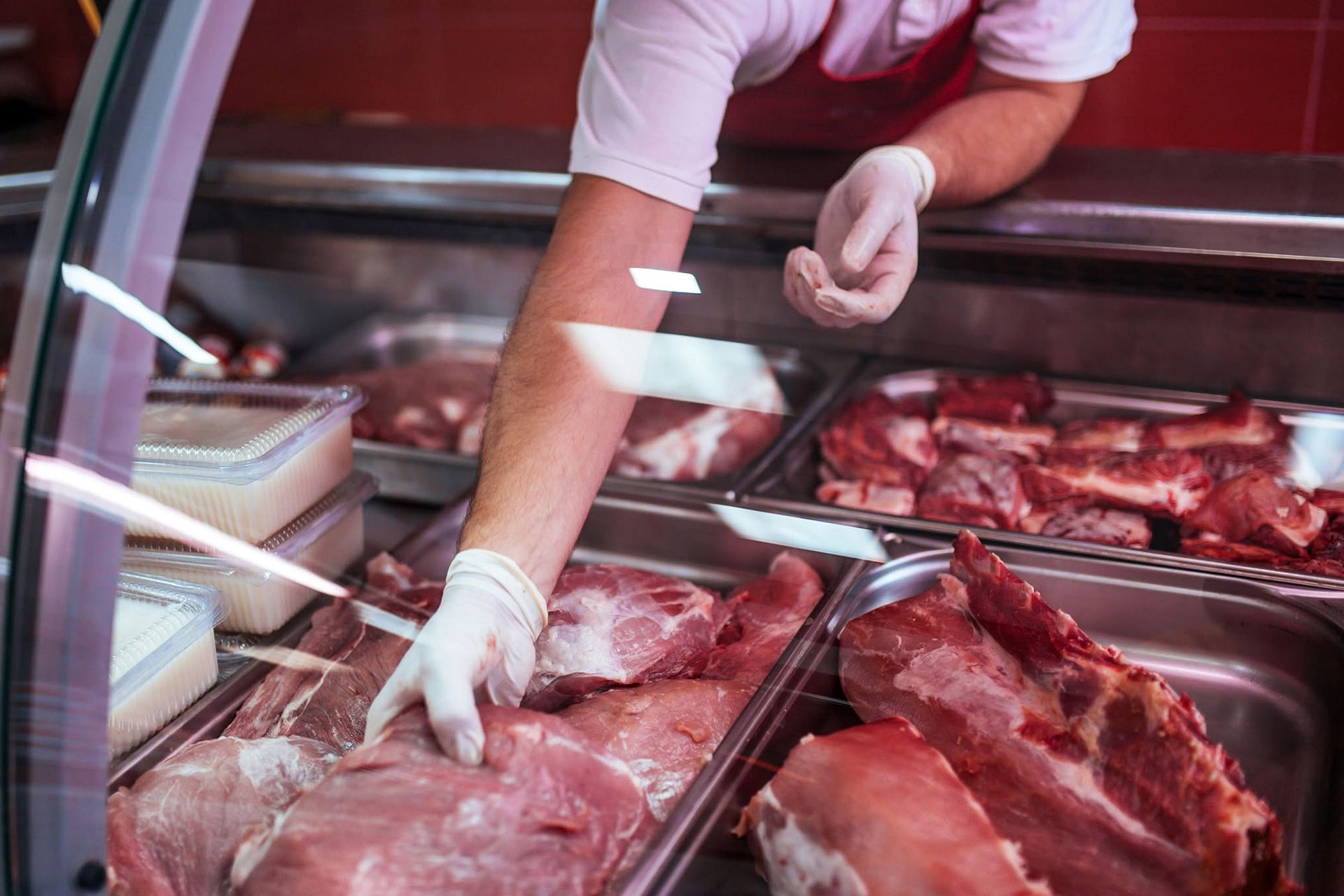 Un macellaio maneggia carne cruda in una vetrina refrigerata, con vassoi di tagli rossi e una mano guantata.