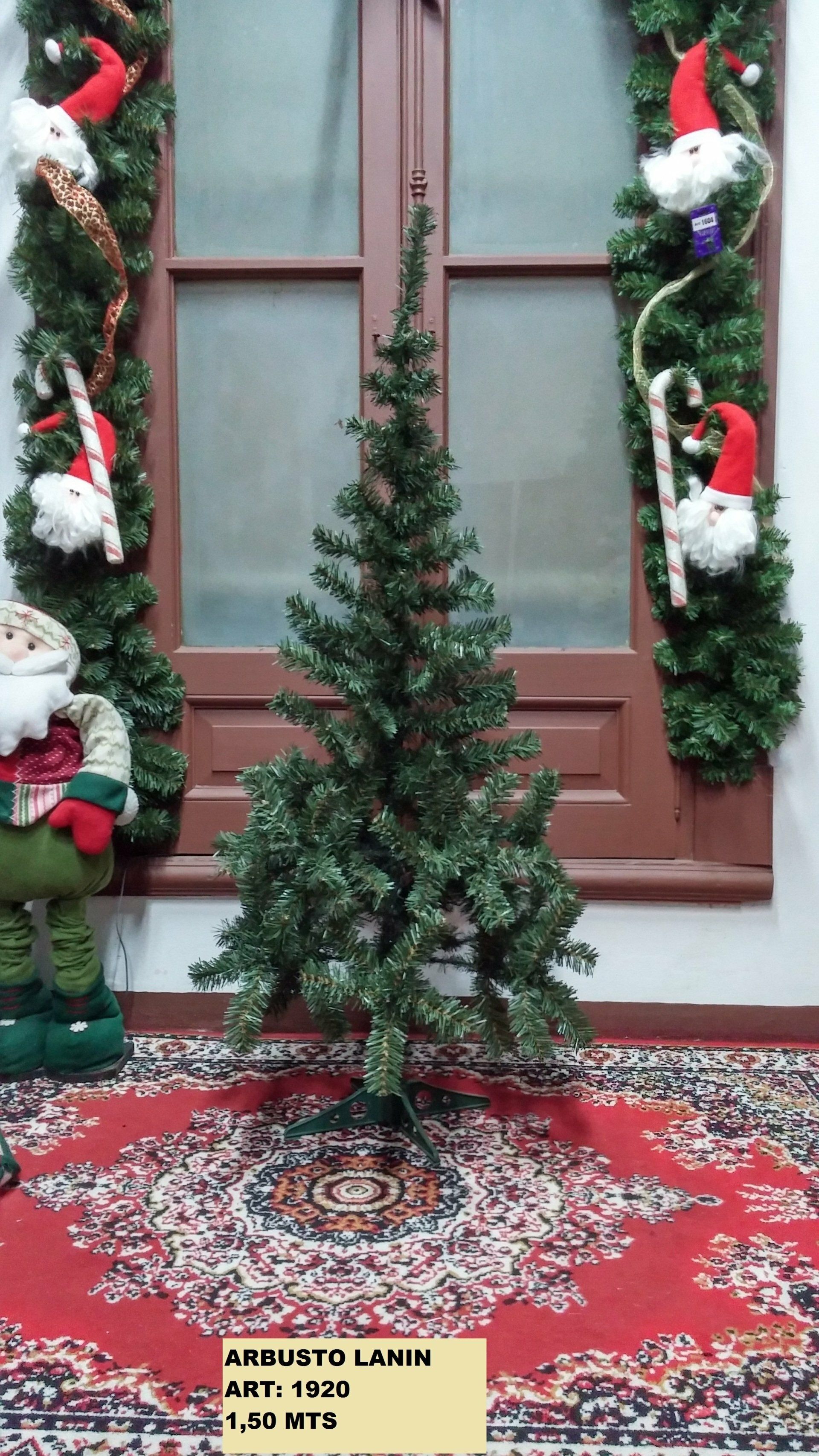 Un pequeño árbol de Navidad desnudo sobre una alfombra estampada entre adornos navideños junto a una puerta marrón.