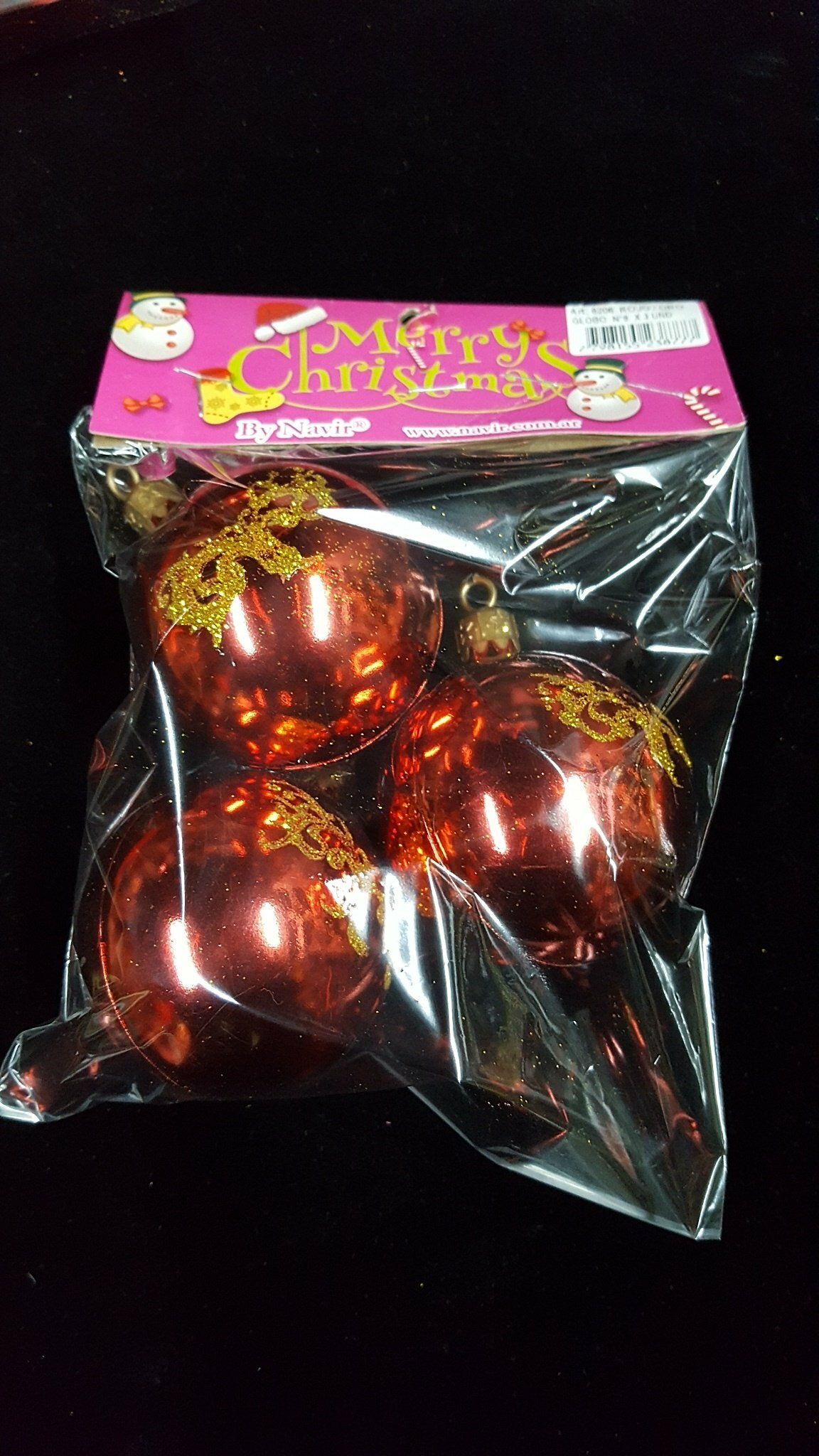 Juego de adornos navideños rojos en envase de plástico transparente con etiqueta rosa sobre fondo negro.