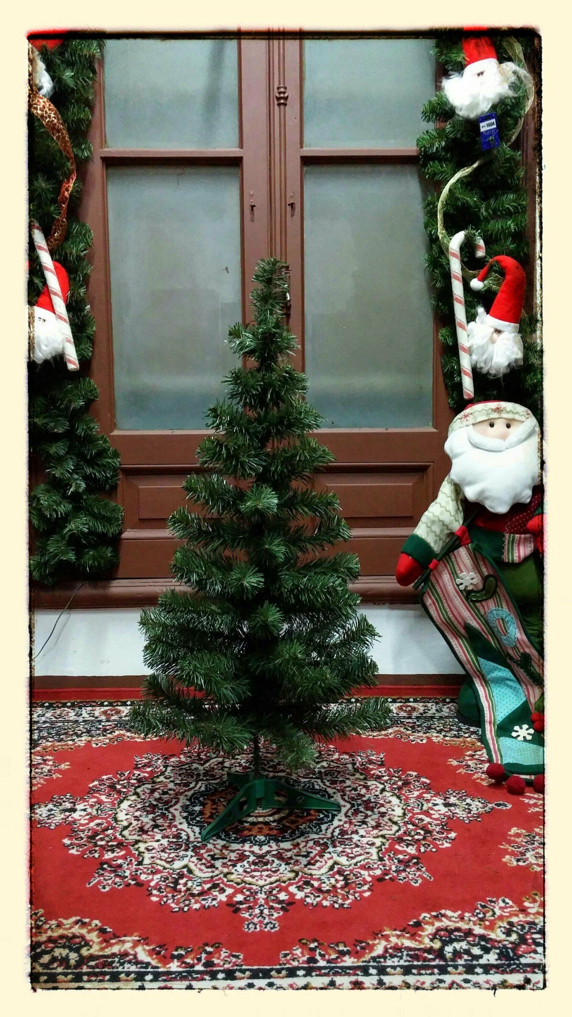 Un pequeño árbol de Navidad decorado sobre una alfombra roja junto a una ventana, rodeado de adornos navideños.
