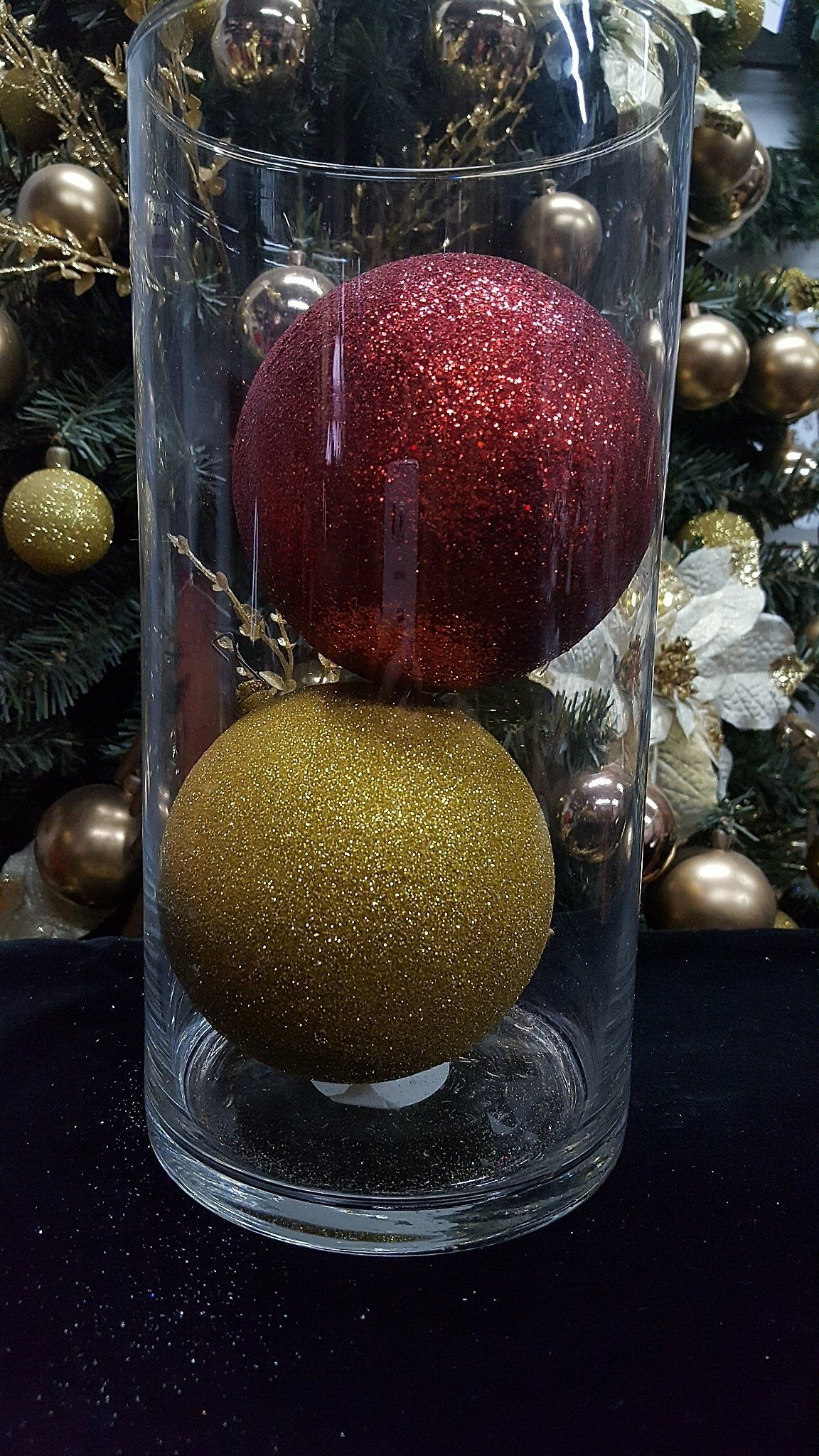 Jarrón cilíndrico transparente con un adorno rojo brillante sobre un adorno dorado, rodeado de decoraciones navideñas.