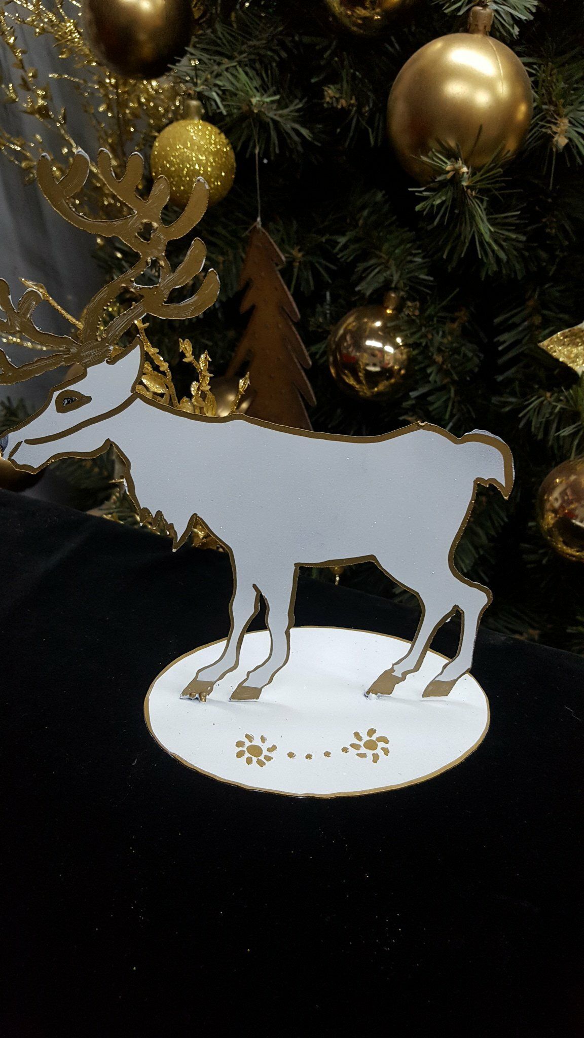 Silueta de ciervo blanco sobre una base nevada, como adorno, frente a un árbol de Navidad decorado.