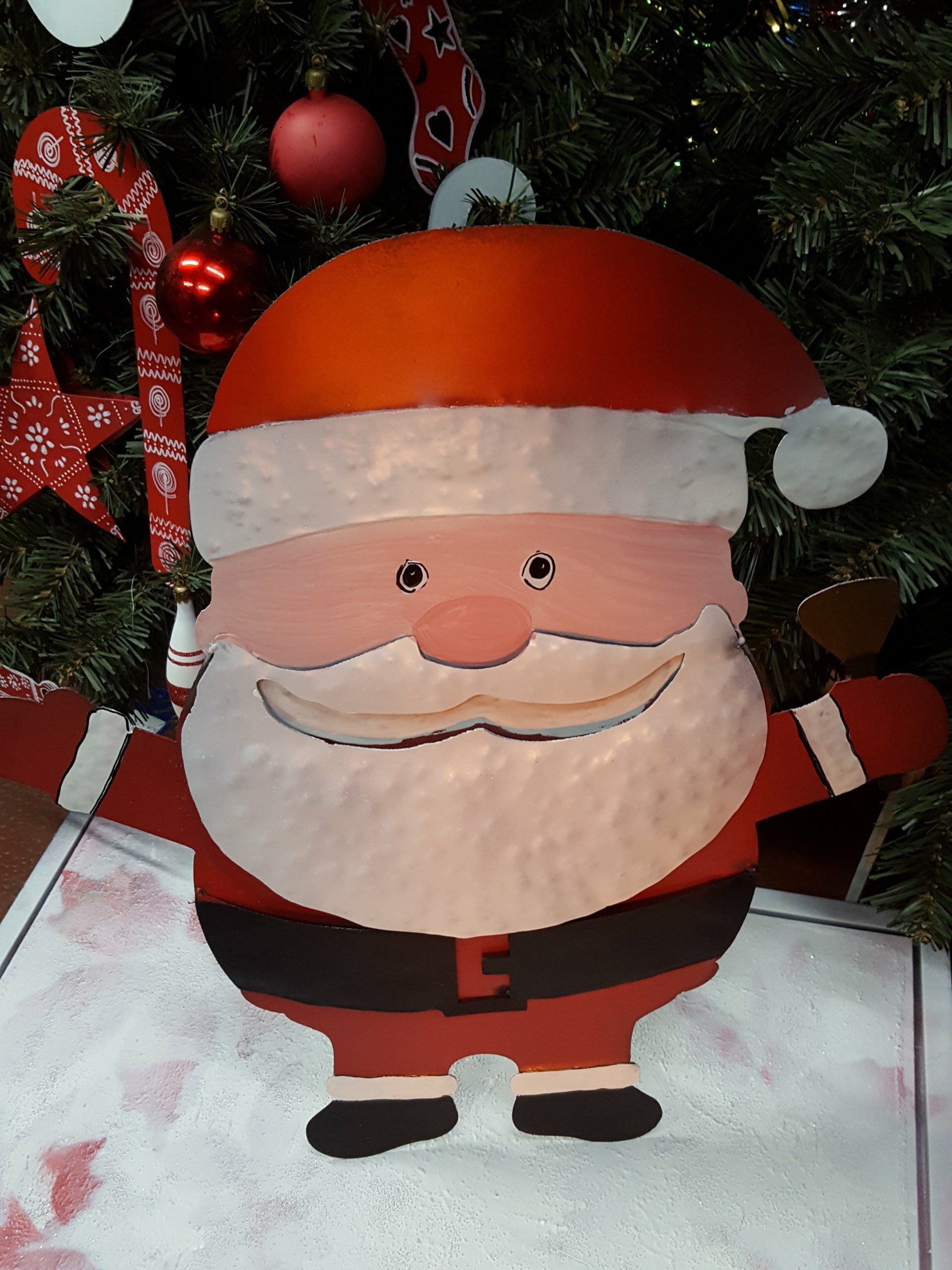 Adorno de Papá Noel sonriente con gorro rojo y cinturón negro, de pie frente a un árbol de Navidad.