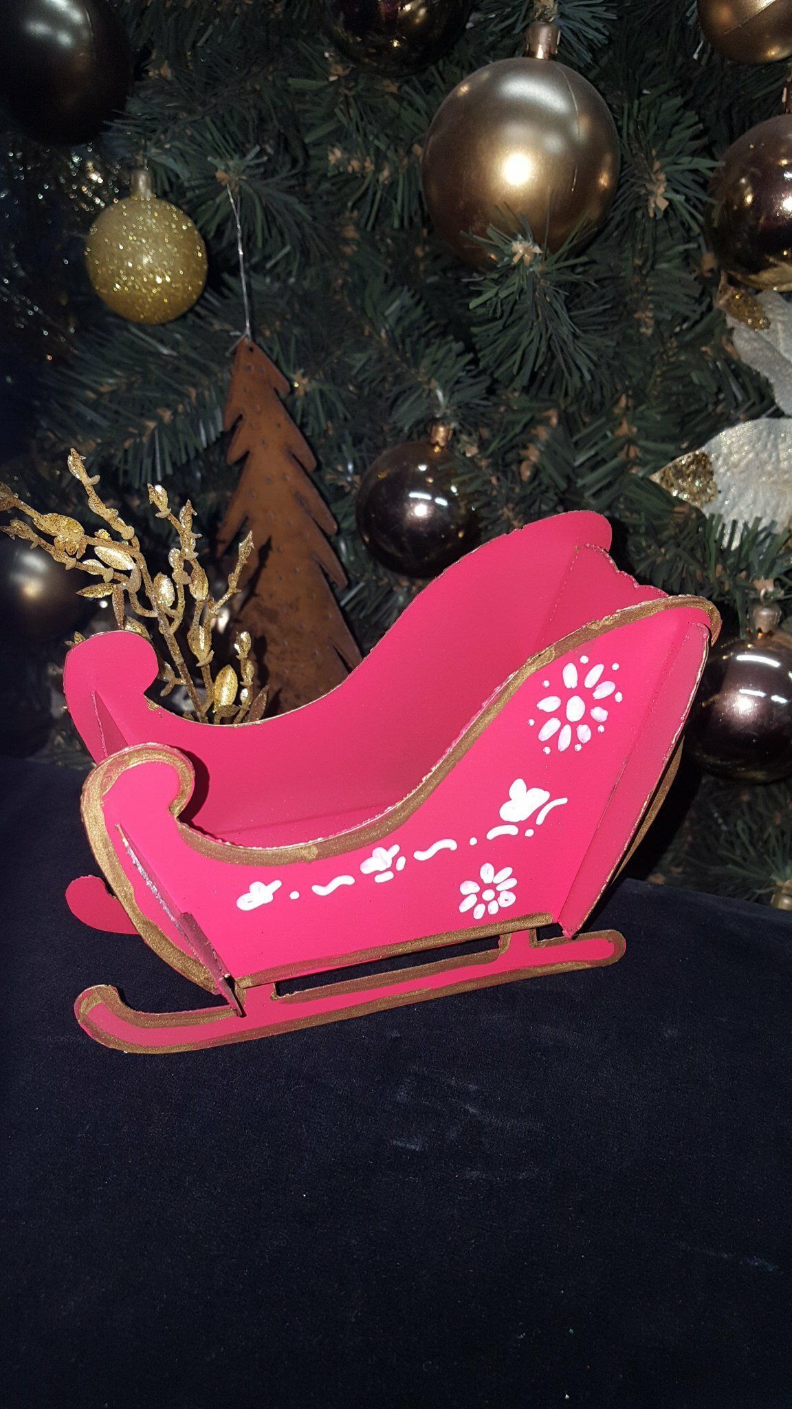 Trineo decorativo rosa delante de un árbol de Navidad con bolas doradas y plateadas.