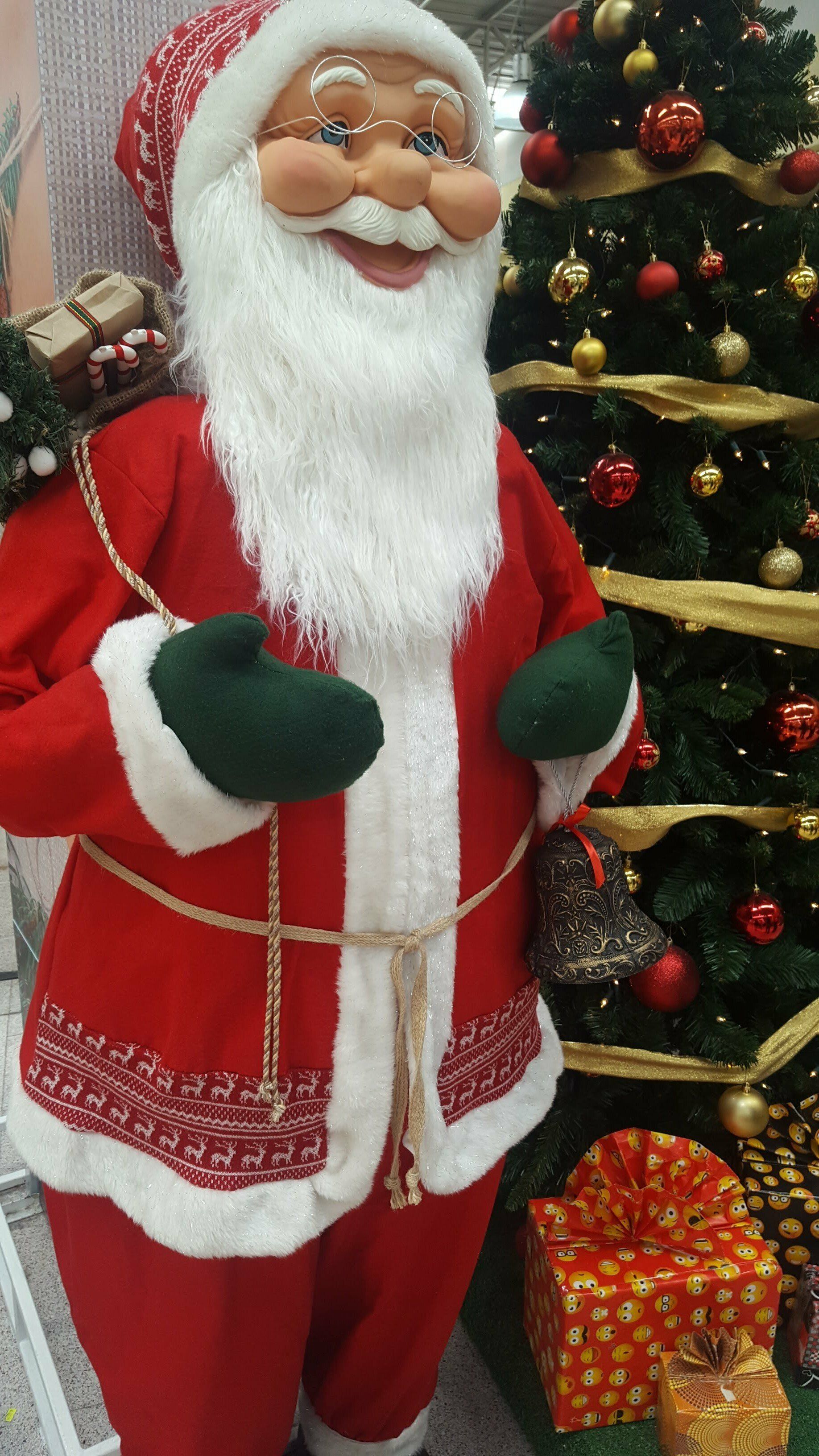 Figura de Papá Noel con traje rojo y barba blanca de pie junto a un árbol de Navidad decorado.