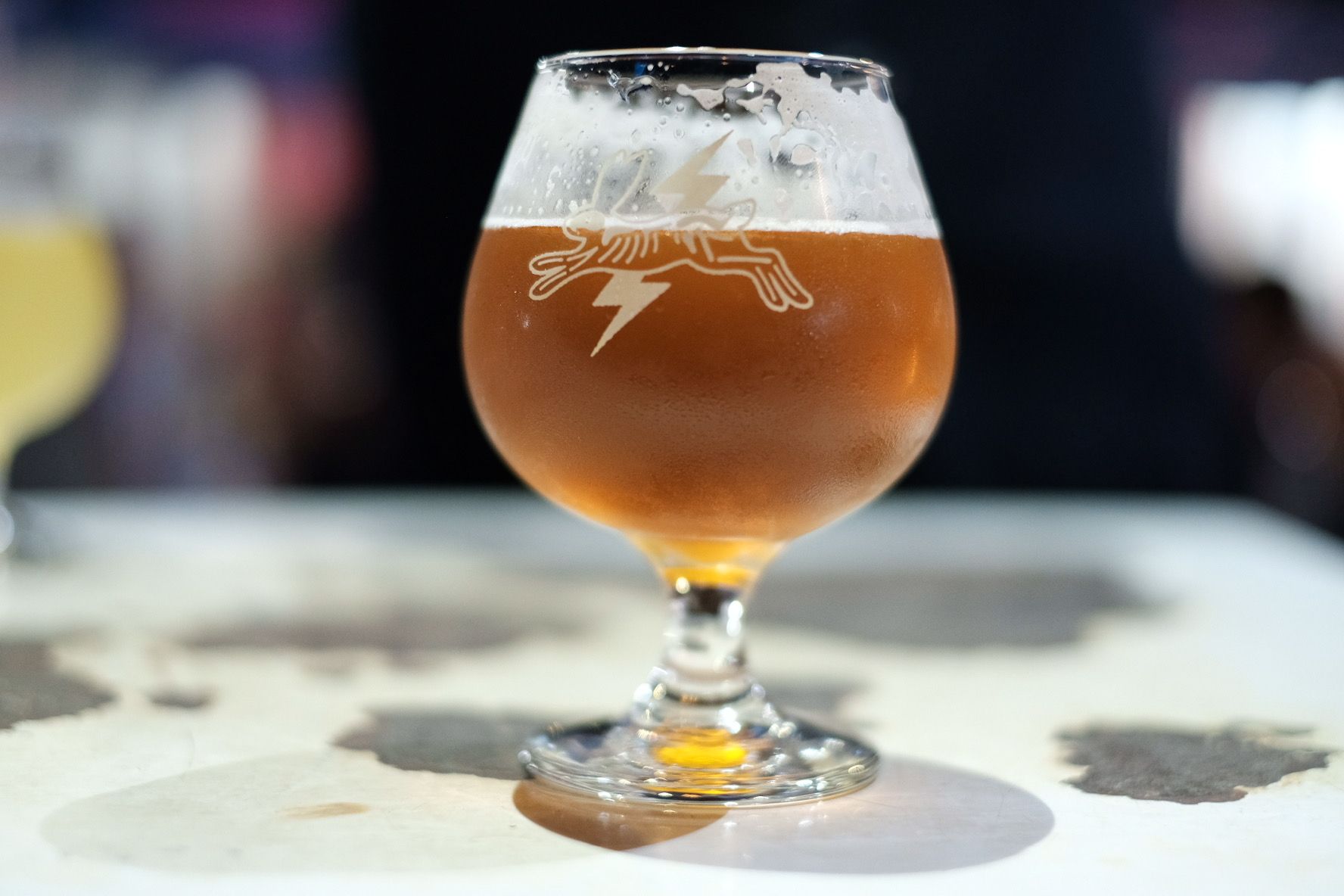 Goblet of amber beer with a logo of a cat and lightning bolt on a white, spotted table.