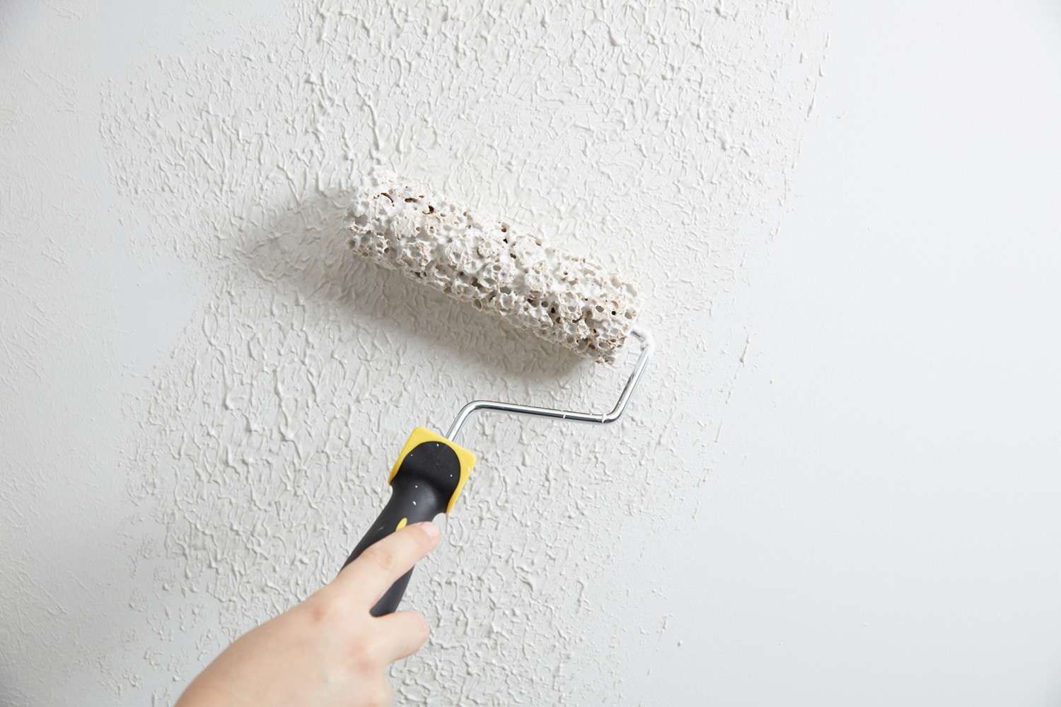 Hand using a paint roller to apply white paint on a textured wall
