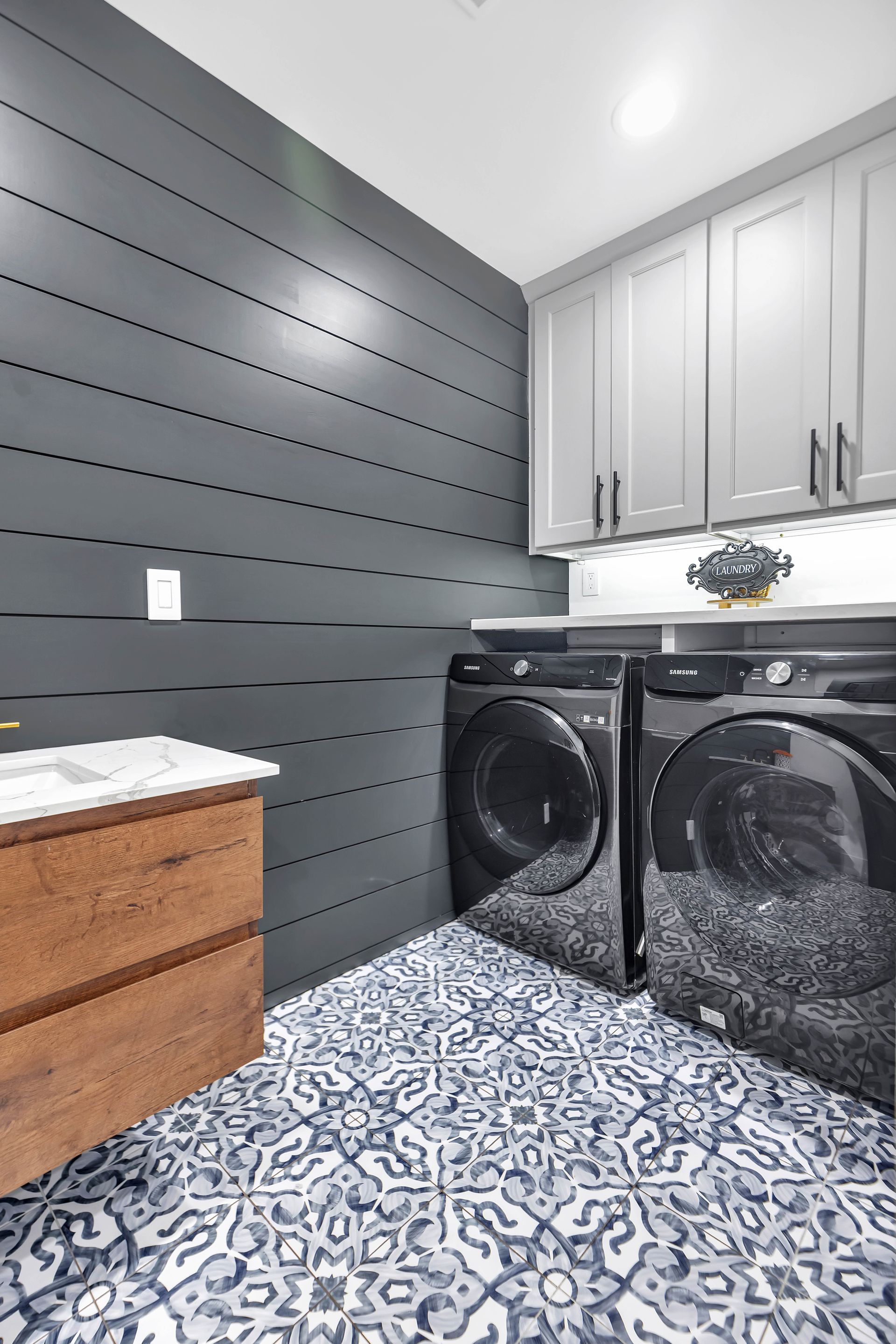 Laundry room with dark gray shiplap wall, black appliances, patterned floor, and gray cabinets.