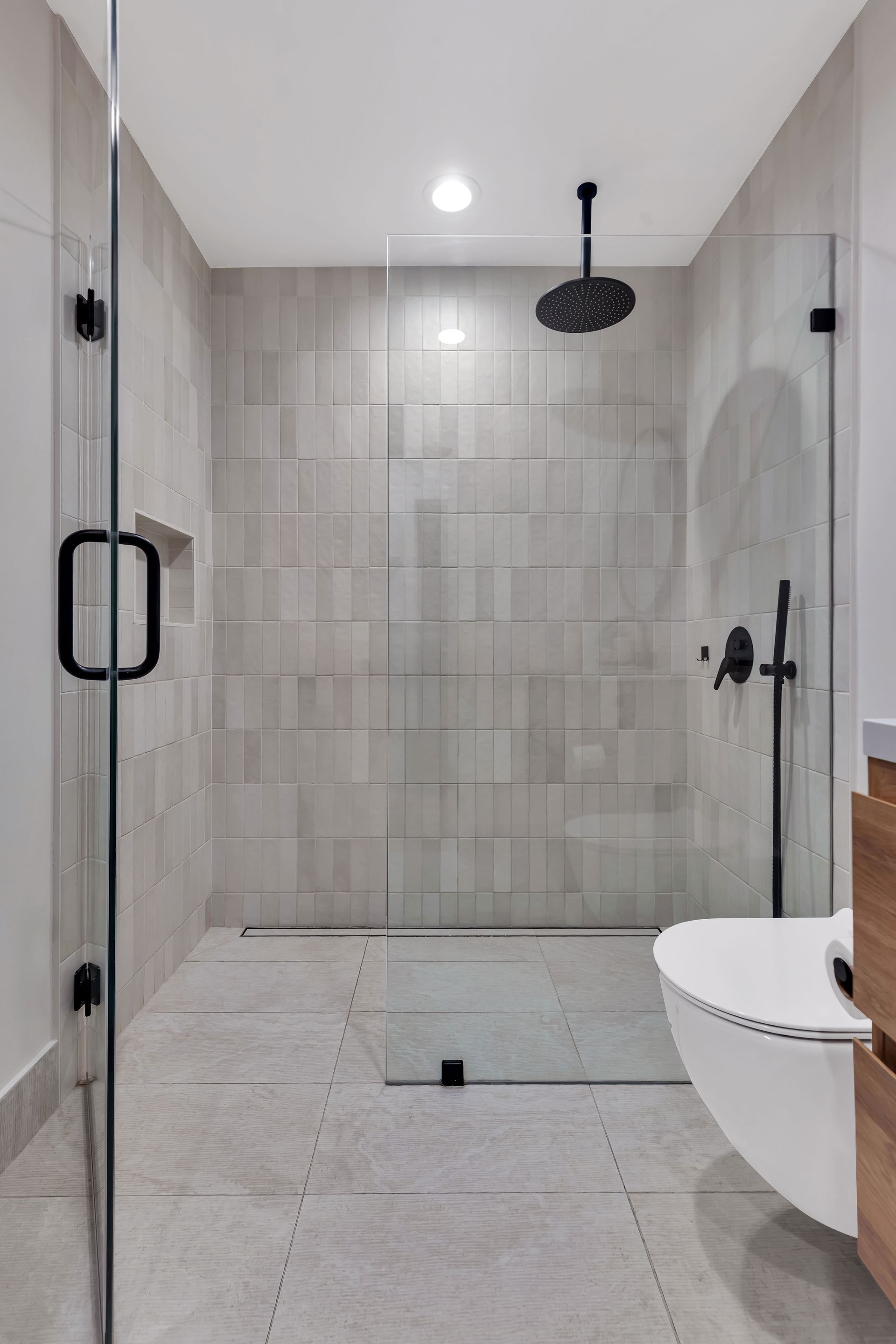 Modern bathroom with gray tiled shower and glass door. Black fixtures and white toilet.