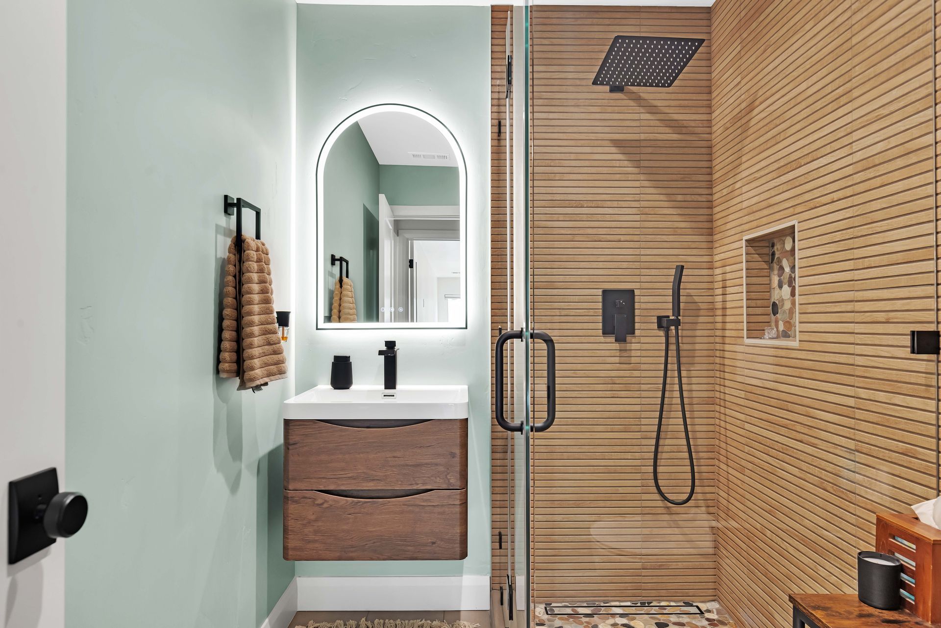 Bathroom with wood-look shower wall, floating vanity, arched mirror with light, and mint green wall.