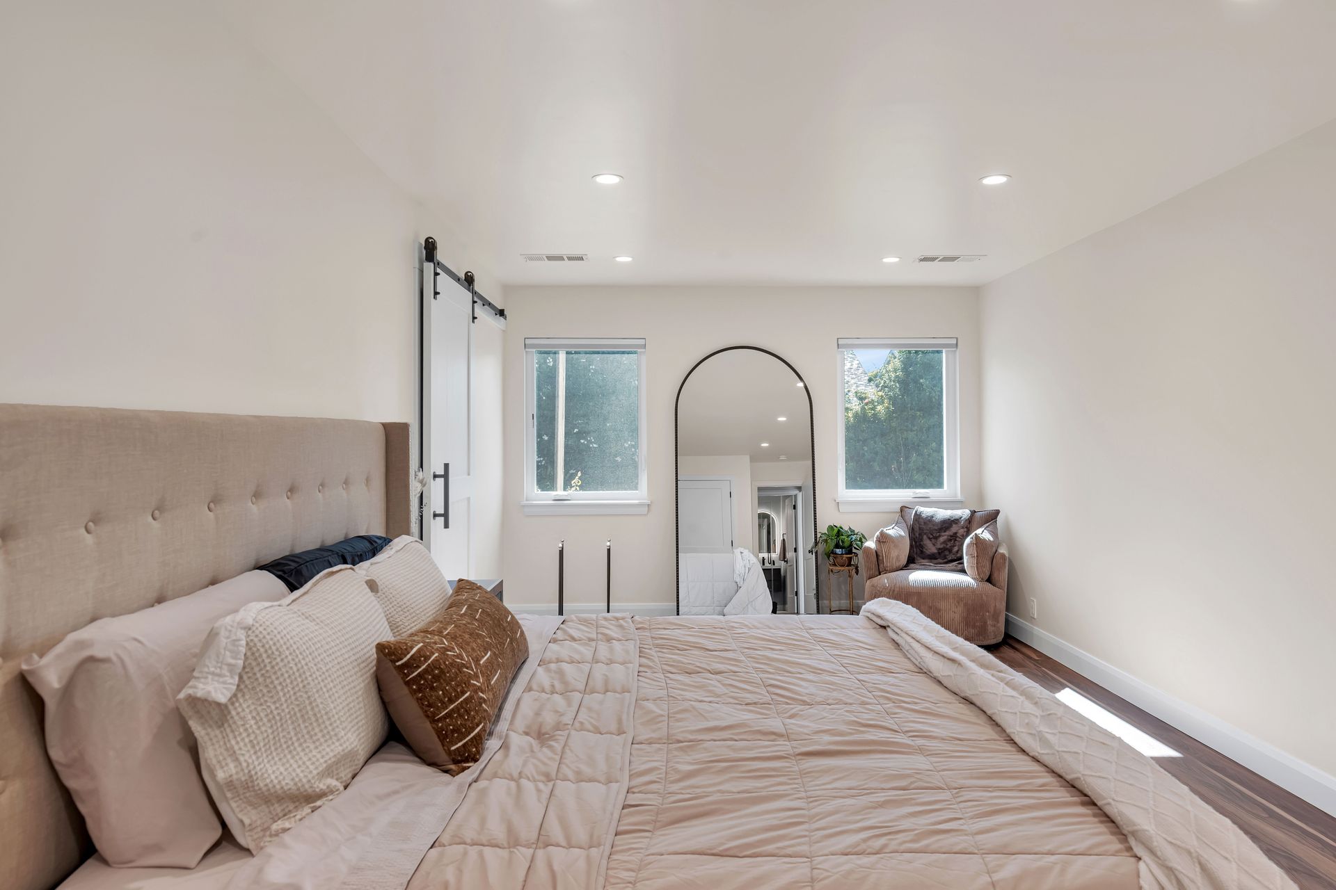 Bedroom with bed, windows, arch mirror, and chair; neutral color palette.