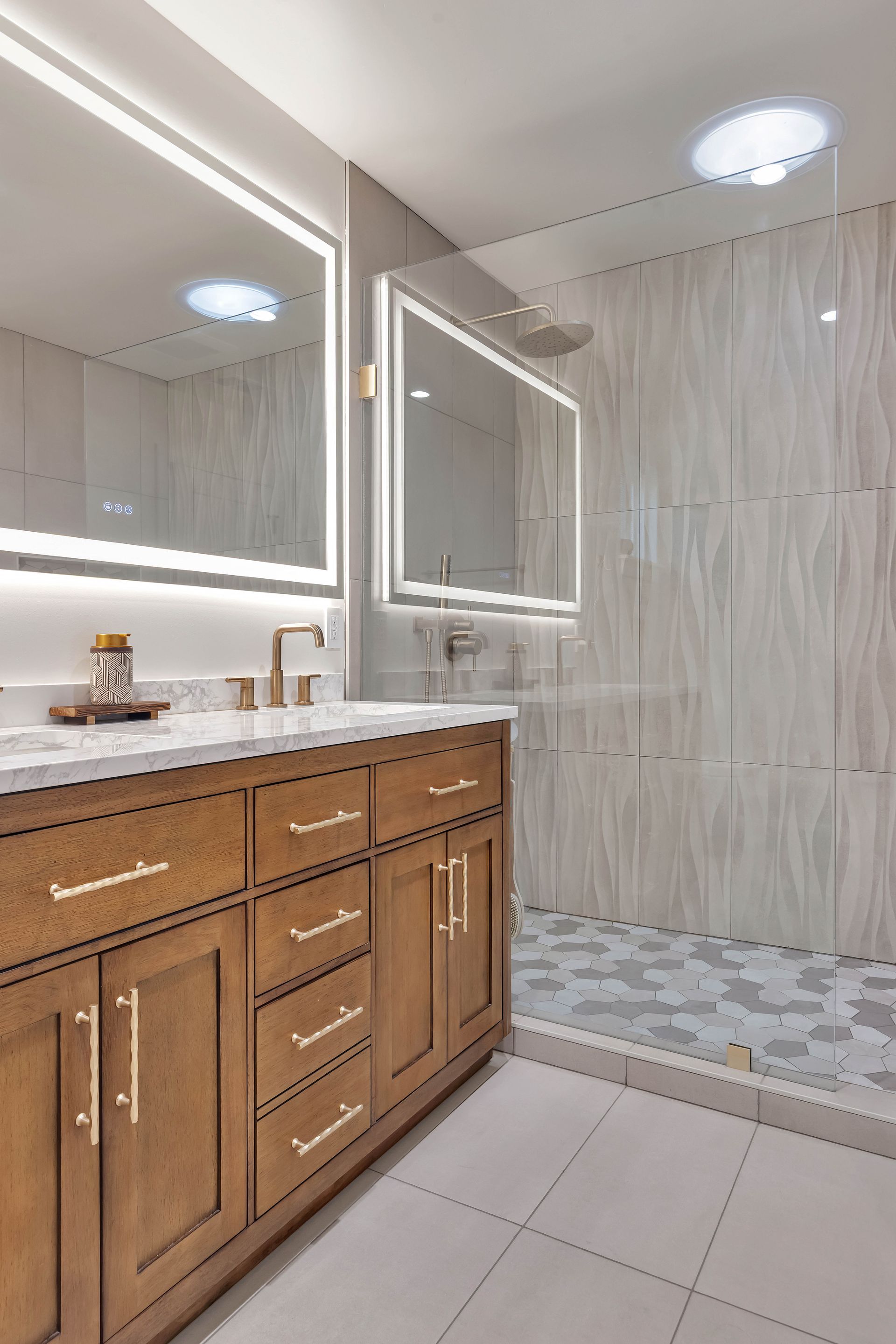 Modern bathroom with light wood vanity, two illuminated mirrors, and a glass shower.