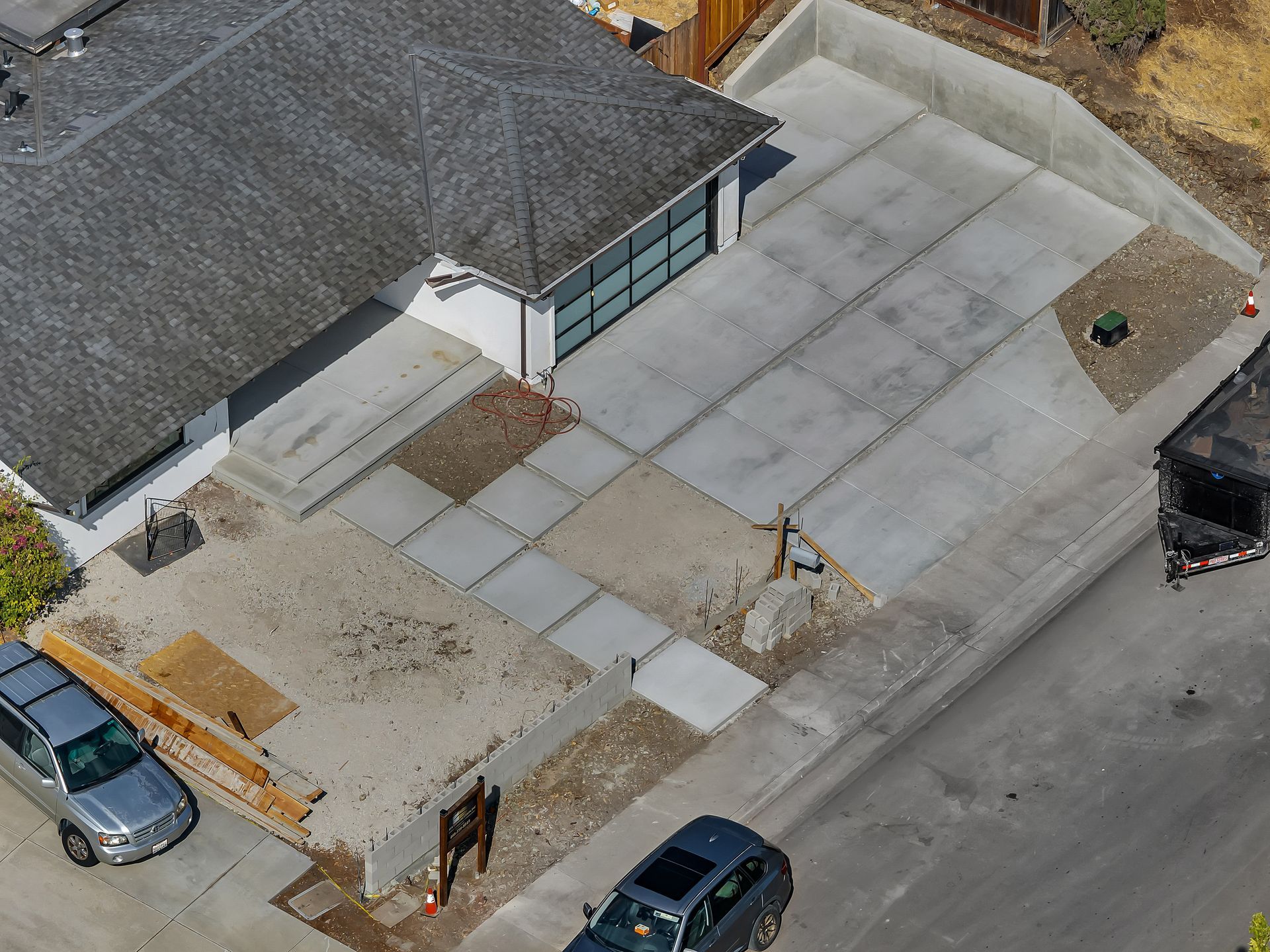 Aerial view of a house with gray roof, concrete driveway, two cars parked on the street. Construction in progress.