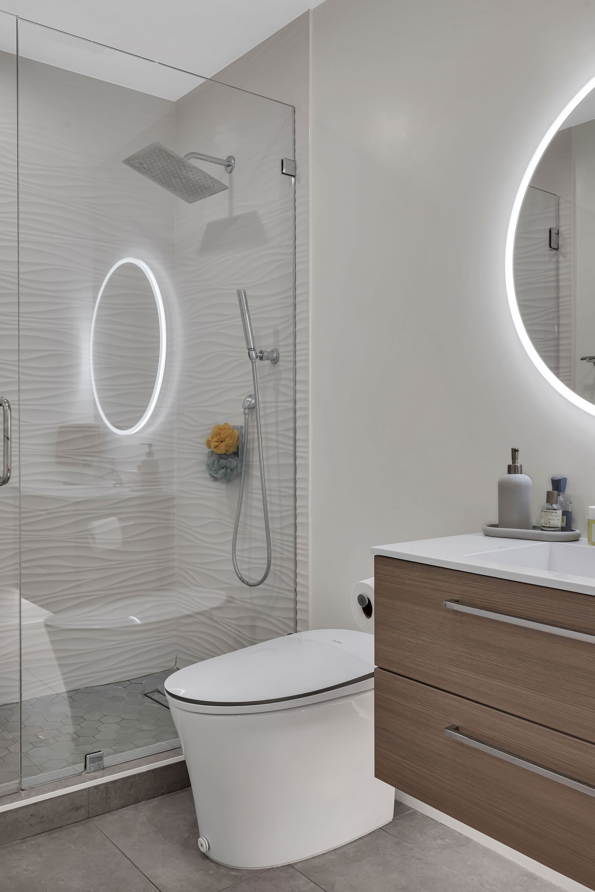 Modern bathroom with glass shower, toilet, floating vanity, and round illuminated mirrors.
