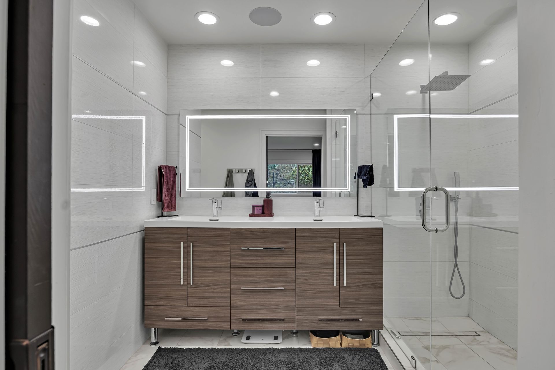 Modern bathroom with brown vanity, white tile, large mirror, and glass shower.