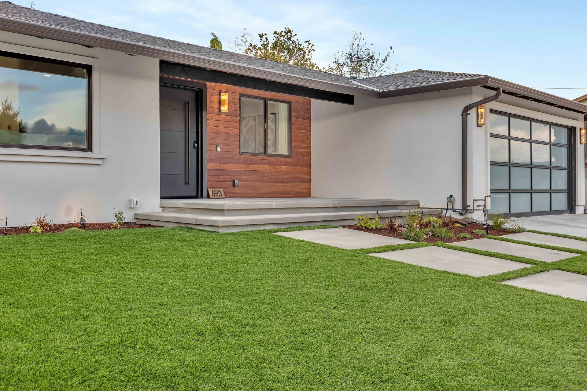 Modern white house with a wood accent entrance, gray roof, and a garage with glass panels. Green lawn.