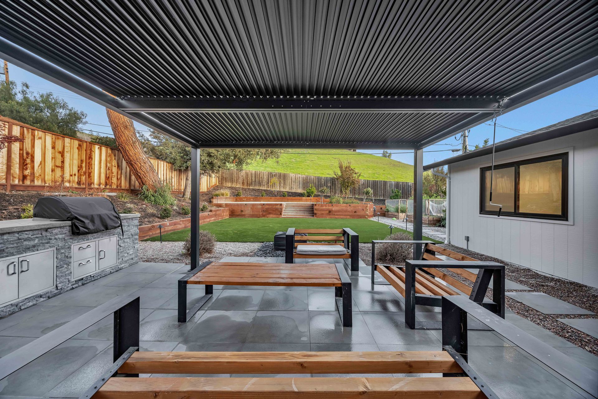 Outdoor patio with grill, seating, and pergola roof overlooking a backyard with lawn and hillside.