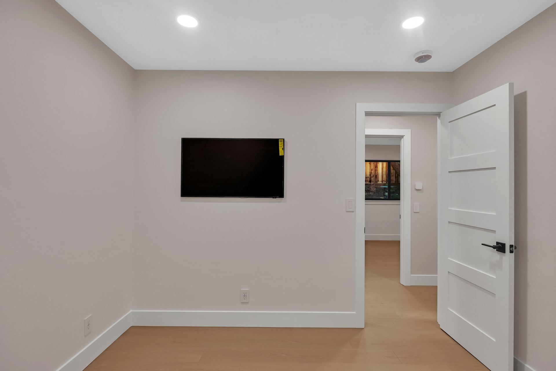 Empty room with a mounted TV, open door, light-colored walls and flooring, recessed lighting.