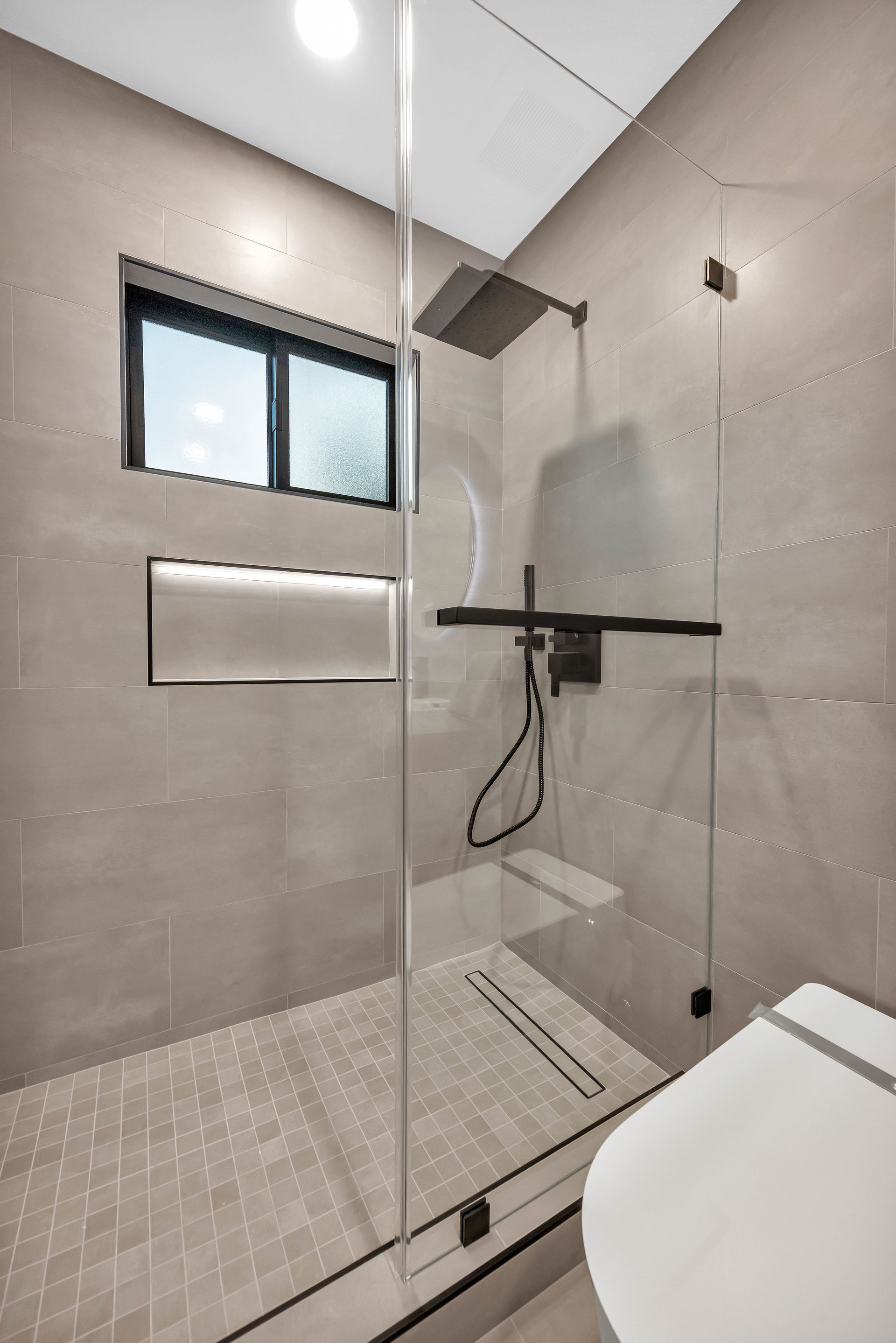 Modern bathroom with glass shower enclosure, gray tiles, and a window.