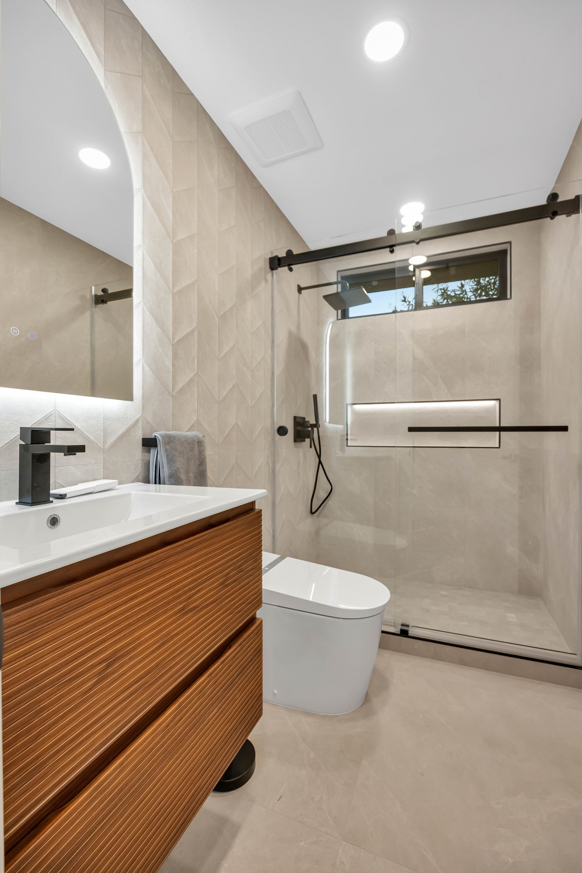 Modern bathroom with wood vanity, white toilet, glass shower, and light beige tiles.