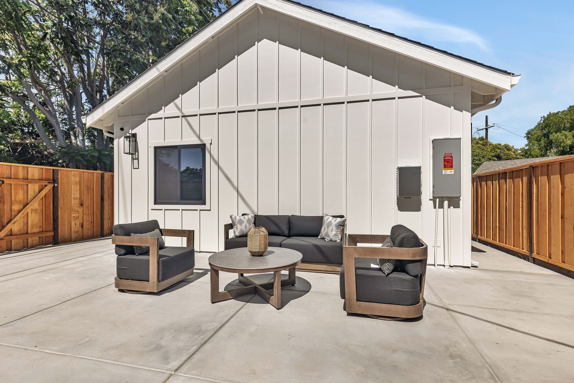 White exterior building with outdoor furniture on a concrete patio, wooden fence, and blue sky.