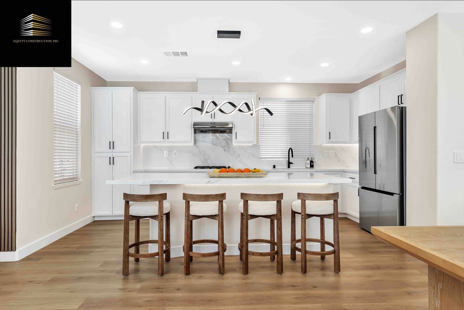 Modern white kitchen with island seating four stools; stainless steel refrigerator.
