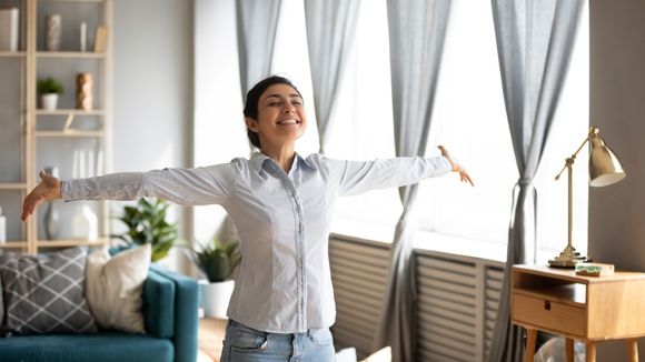 Woman with arms outstretched smiling by a bright window, in a living room.
