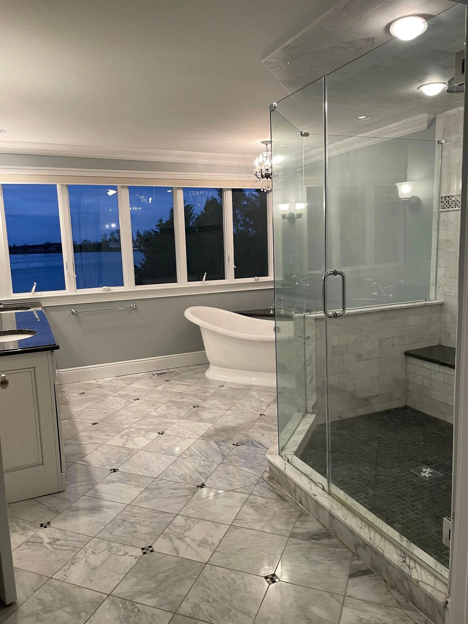 Modern bathroom with glass shower, freestanding tub, and large windows overlooking water at dusk