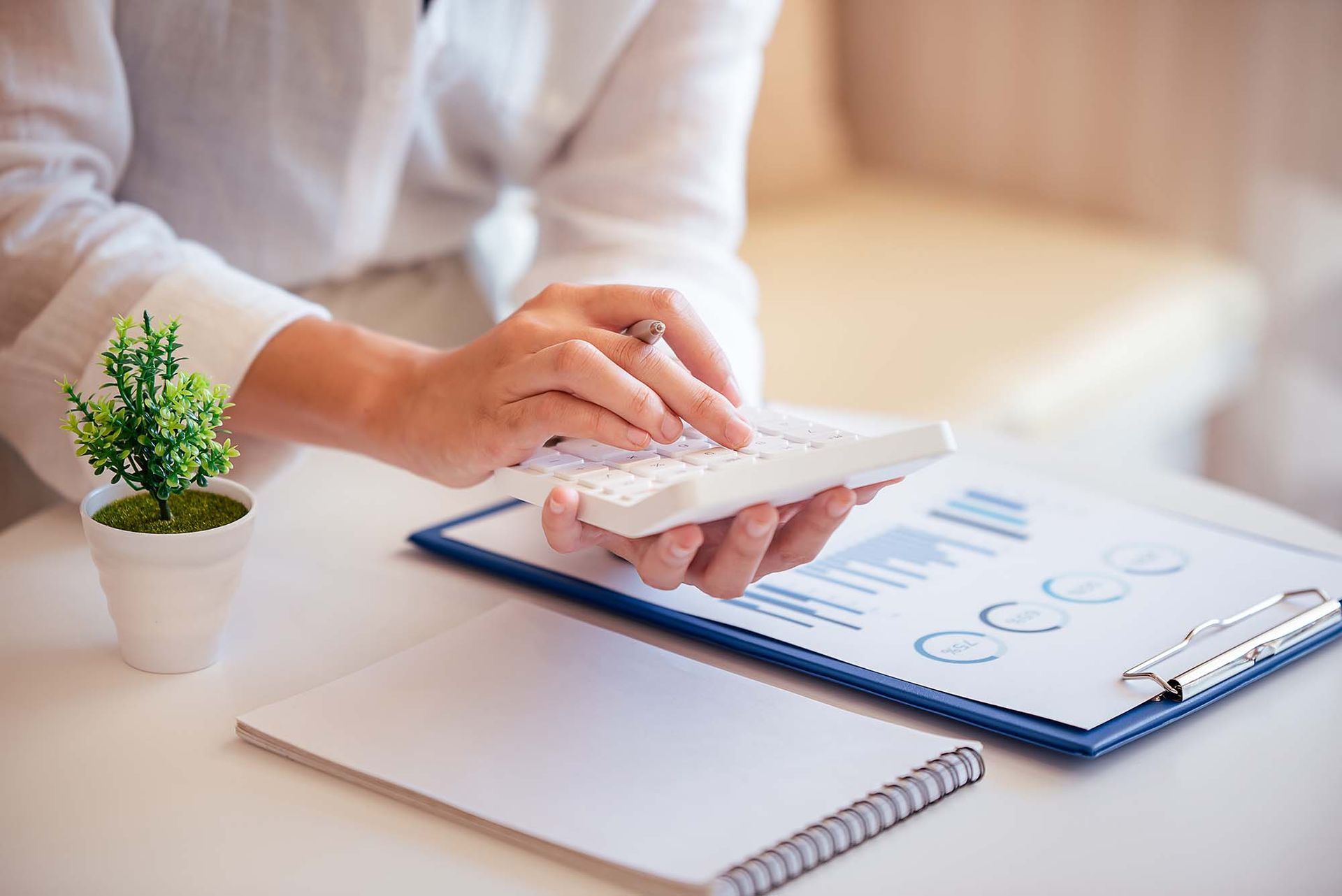 Hands typing on a laptop beside a small potted plant and notebook on a bright desk