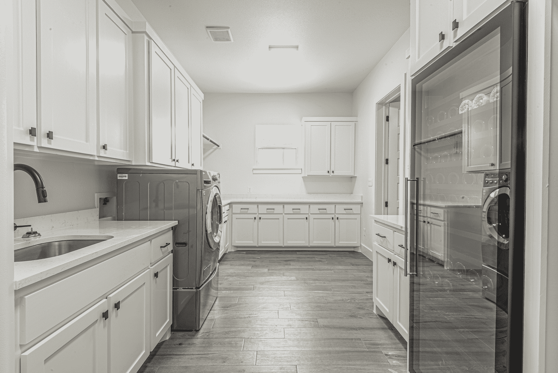 A black and white photo of a laundry room with white cabinets , a sink , and a refrigerator.