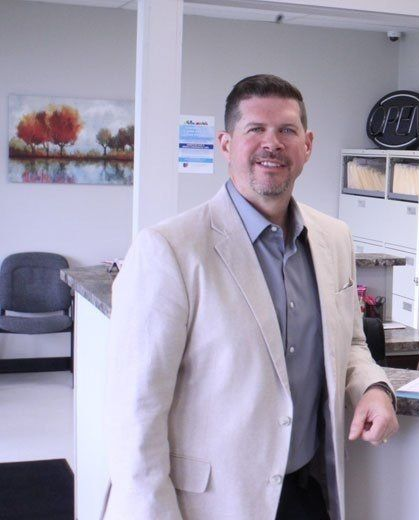 Man in a light blazer smiles, standing behind a reception desk. Interior with artwork and chairs.