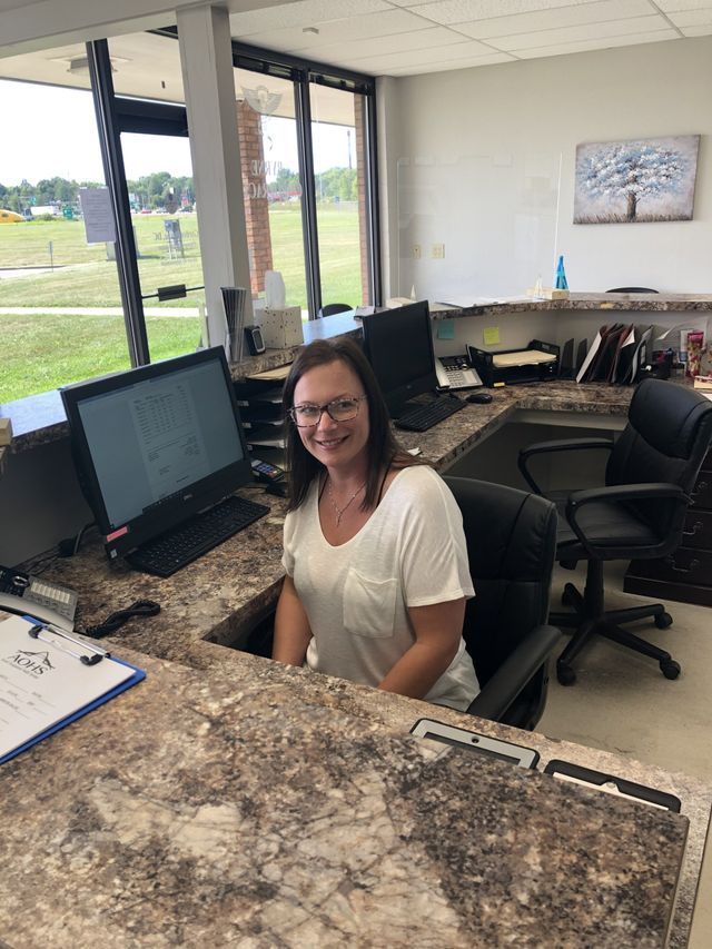 Woman smiling at a reception desk with a computer, in an office setting, with a large window in the background.